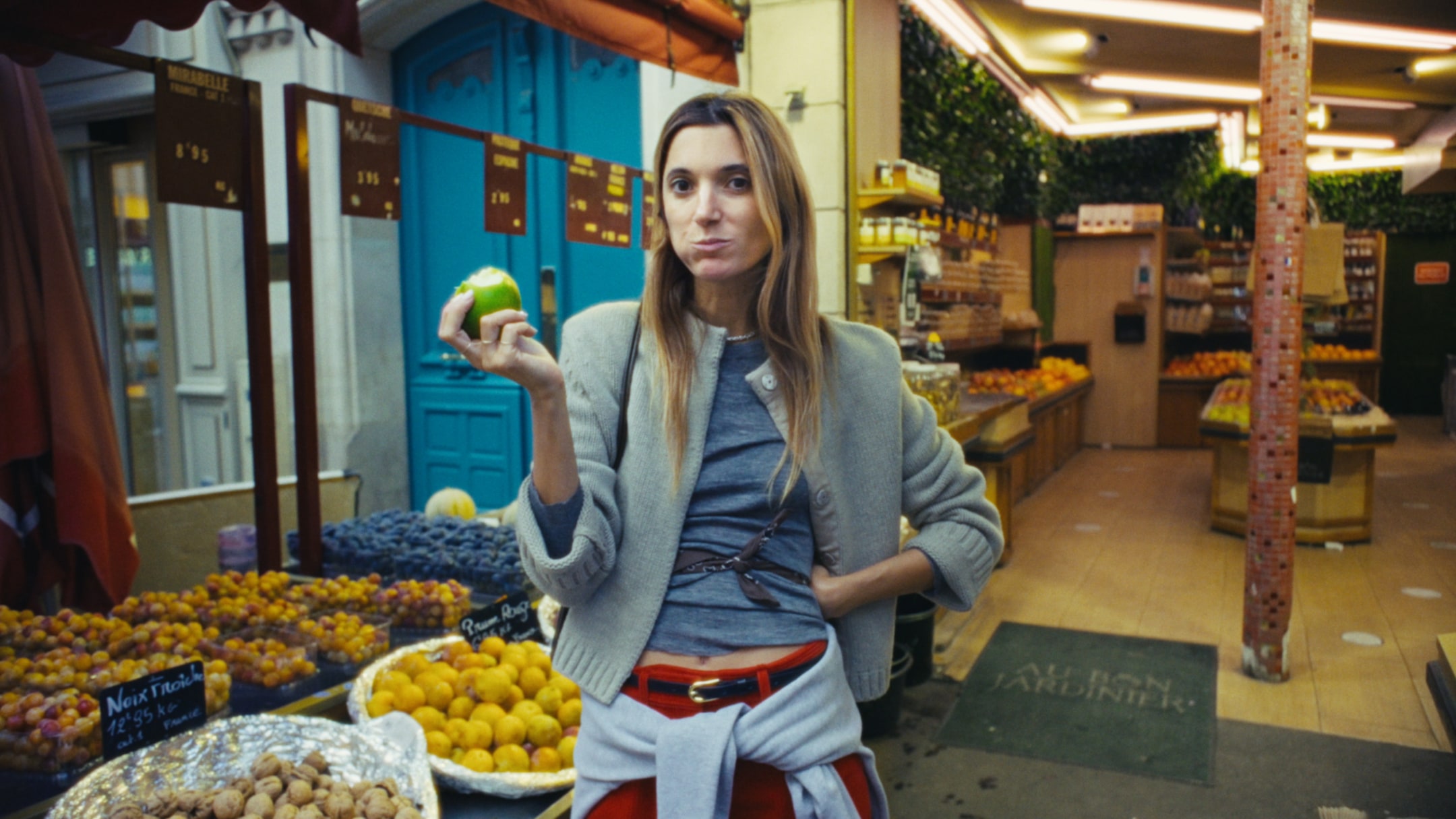 Person standing in a fruit market holds a green apple and appears to be eating it. Various fruits are displayed around them.