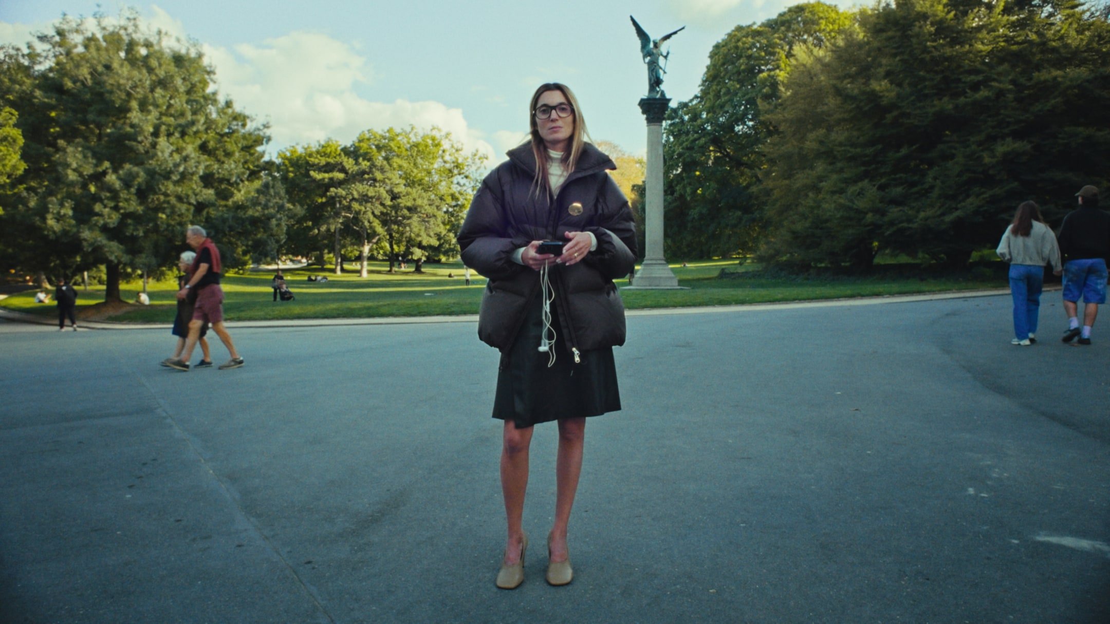 Woman standing in the park wearing a coat, holding a phone and earbuds, with a statue and people walking in the background.