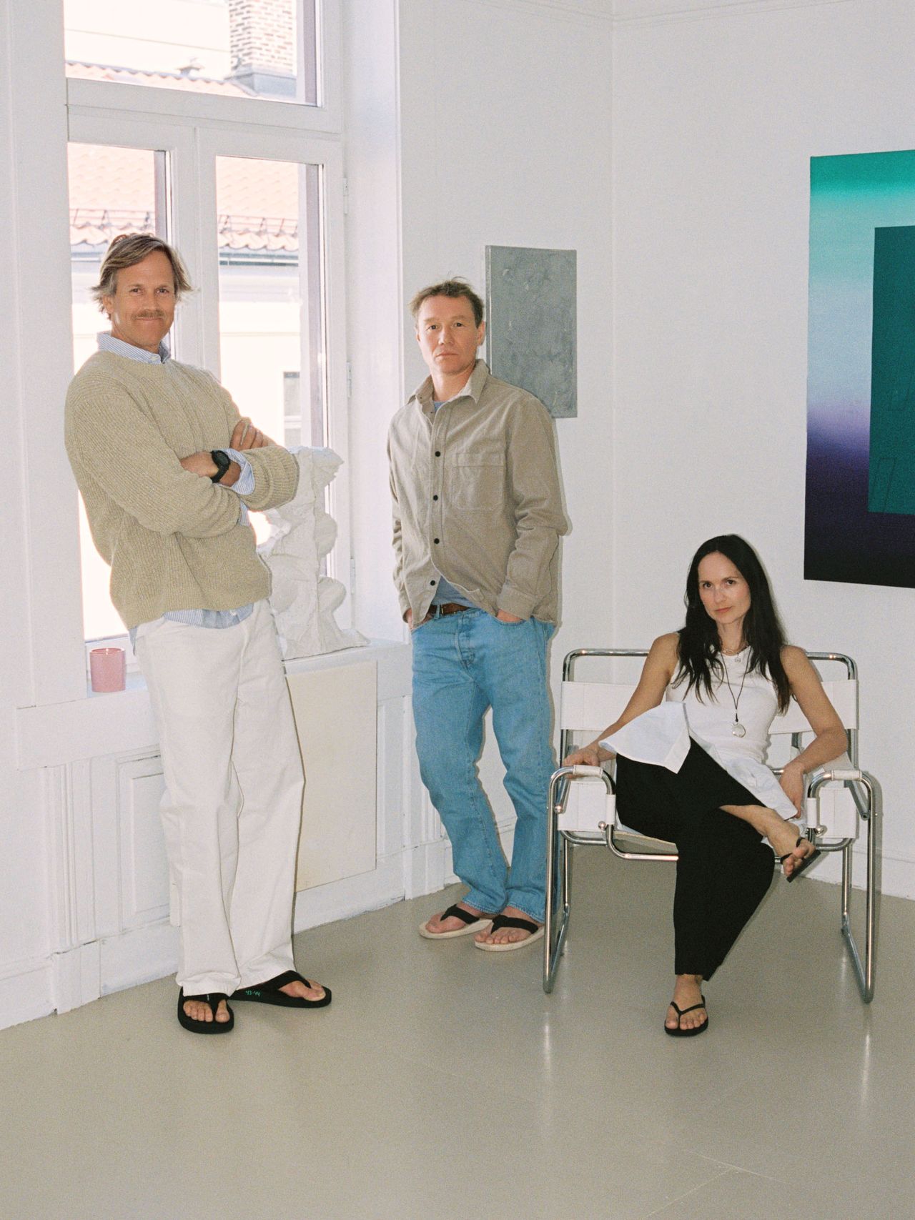 Three people in casual attire pose in a minimalist room with art on the walls and sculptural decor, near a large window.