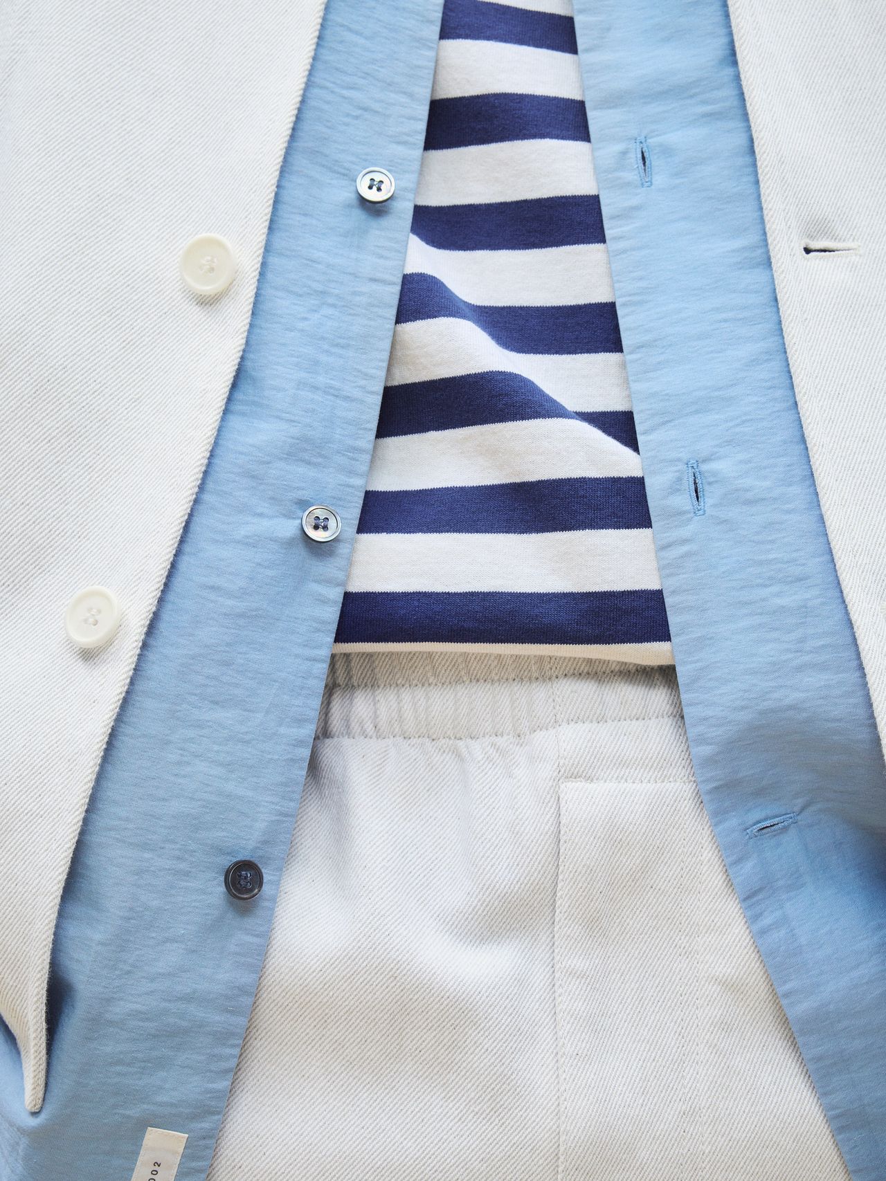 Close-up of a light blazer over a blue and white striped shirt and blue undershirt, paired with light-colored pants.