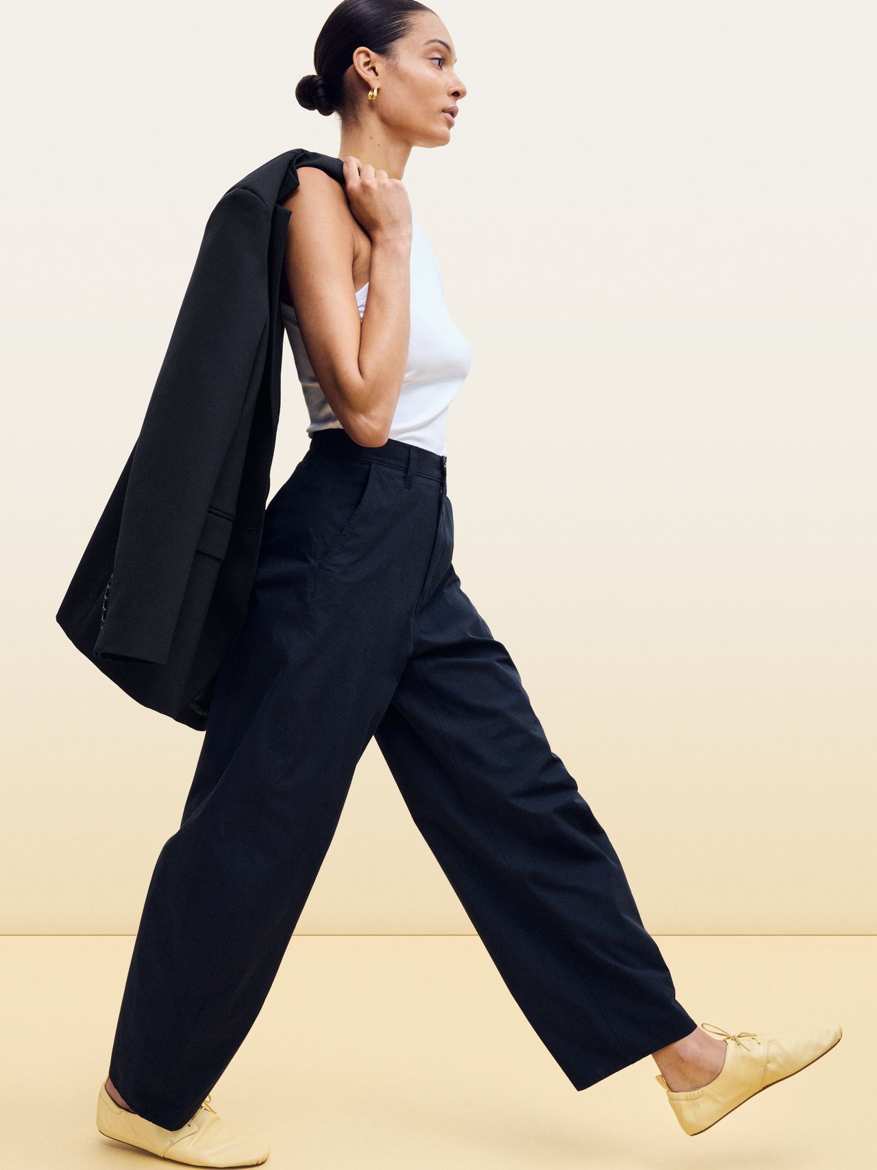 Woman in white sleeveless top, black trousers, and yellow shoes, carries a dark jacket over her shoulder, walking against a beige background.