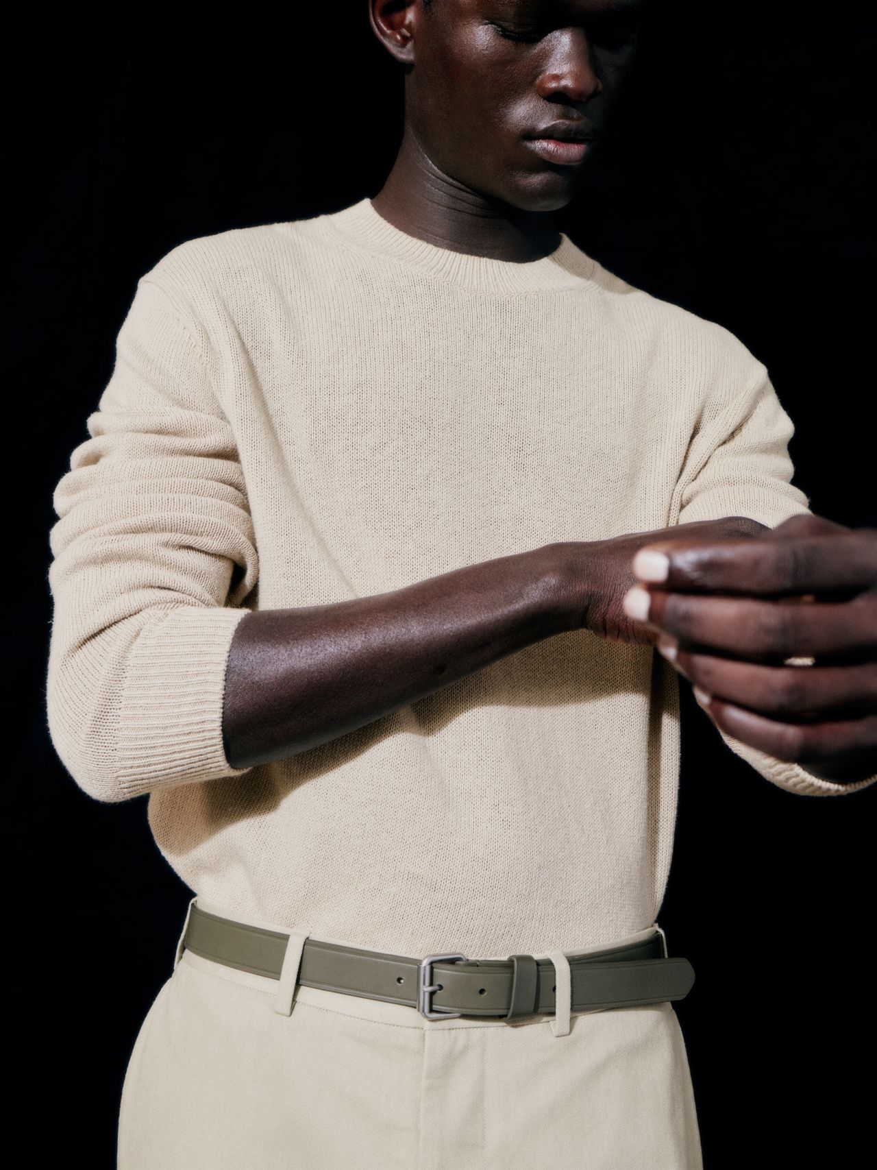 Person in a beige sweater adjusting their sleeve, wearing matching pants and a green belt, against a dark background.