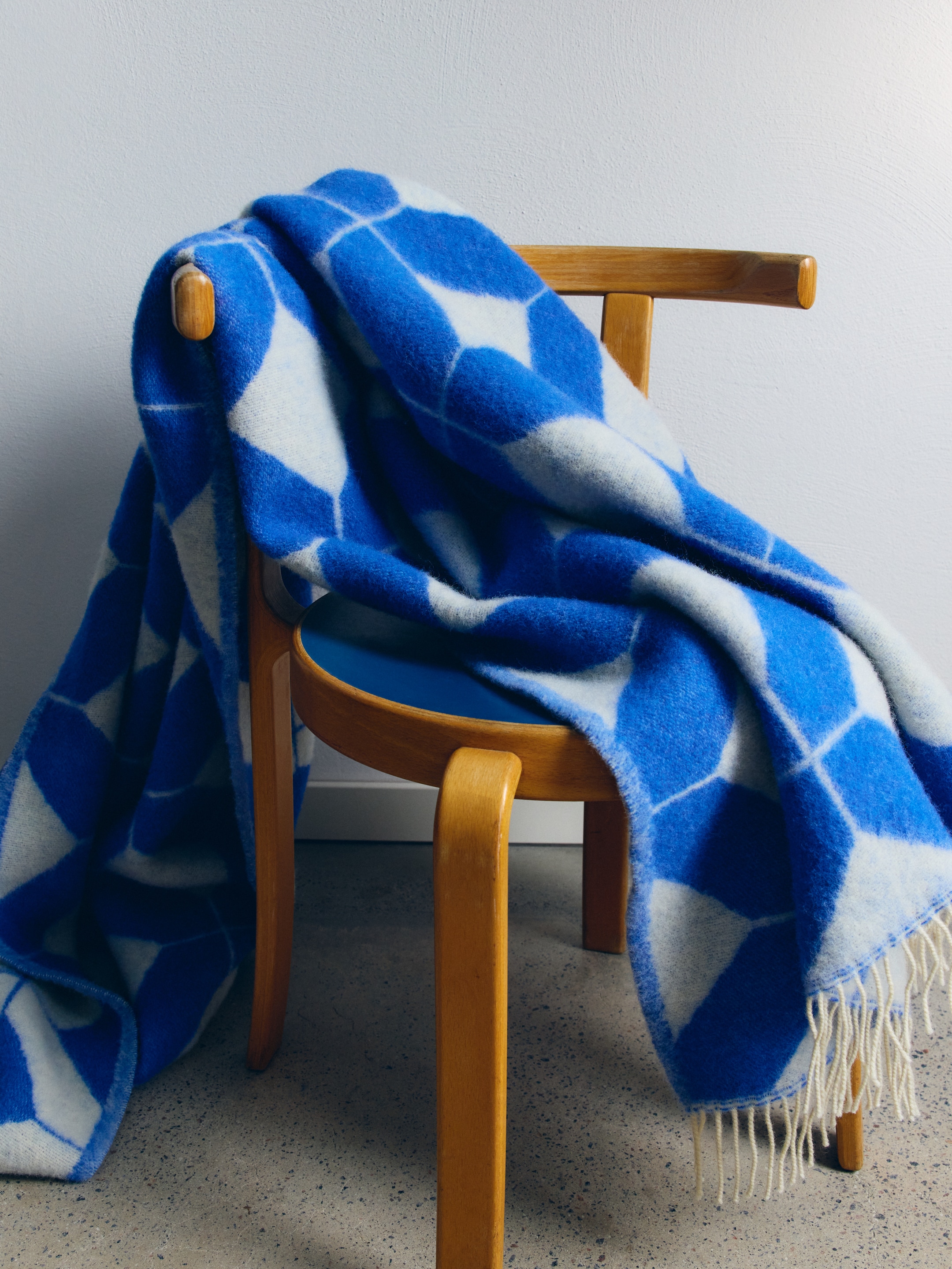 A blue and white geometric-patterned blanket draped over a wooden chair with a blue seat, against a light gray wall.