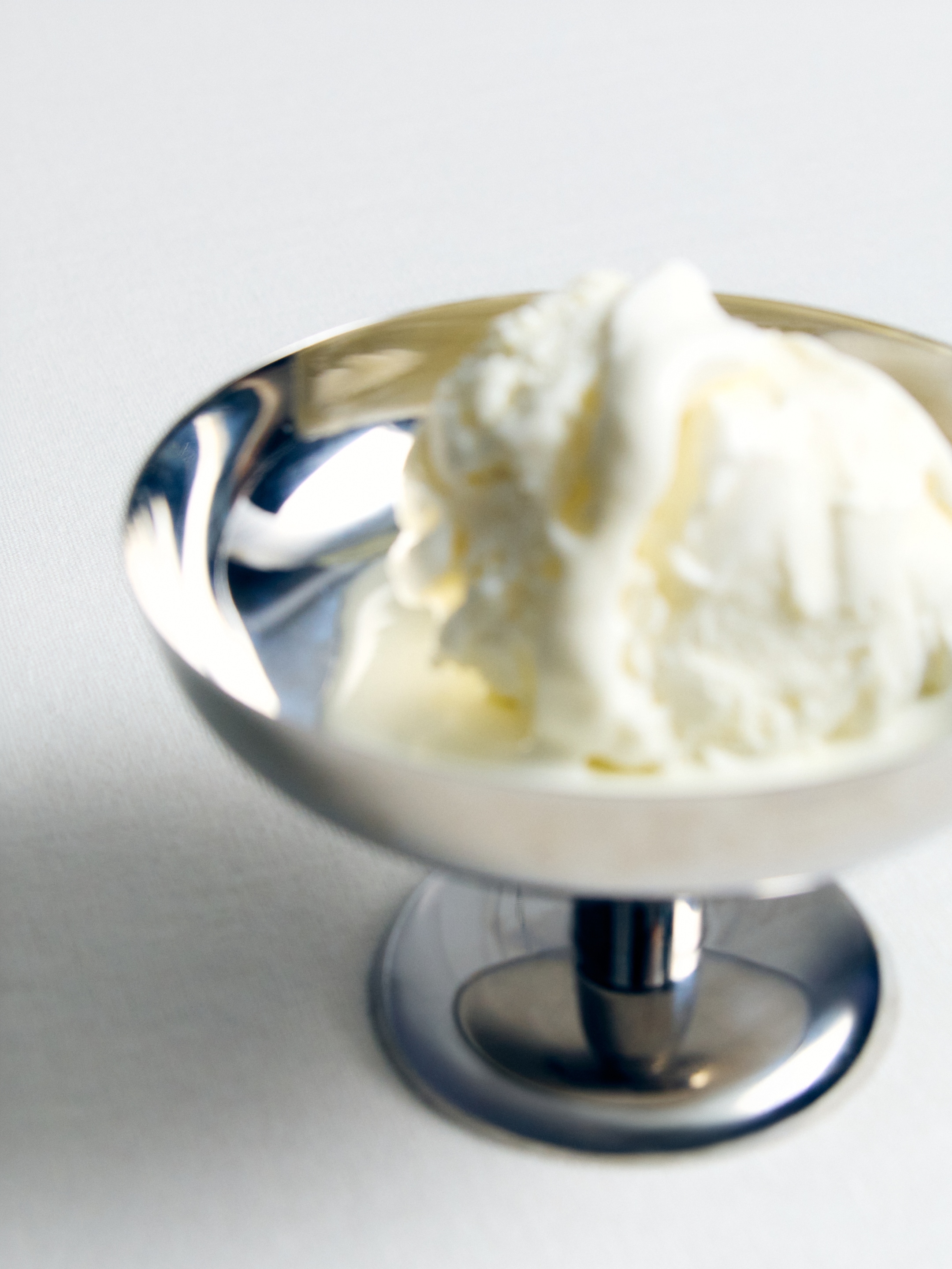 A scoop of vanilla ice cream in a shiny metal dessert cup on a white surface.