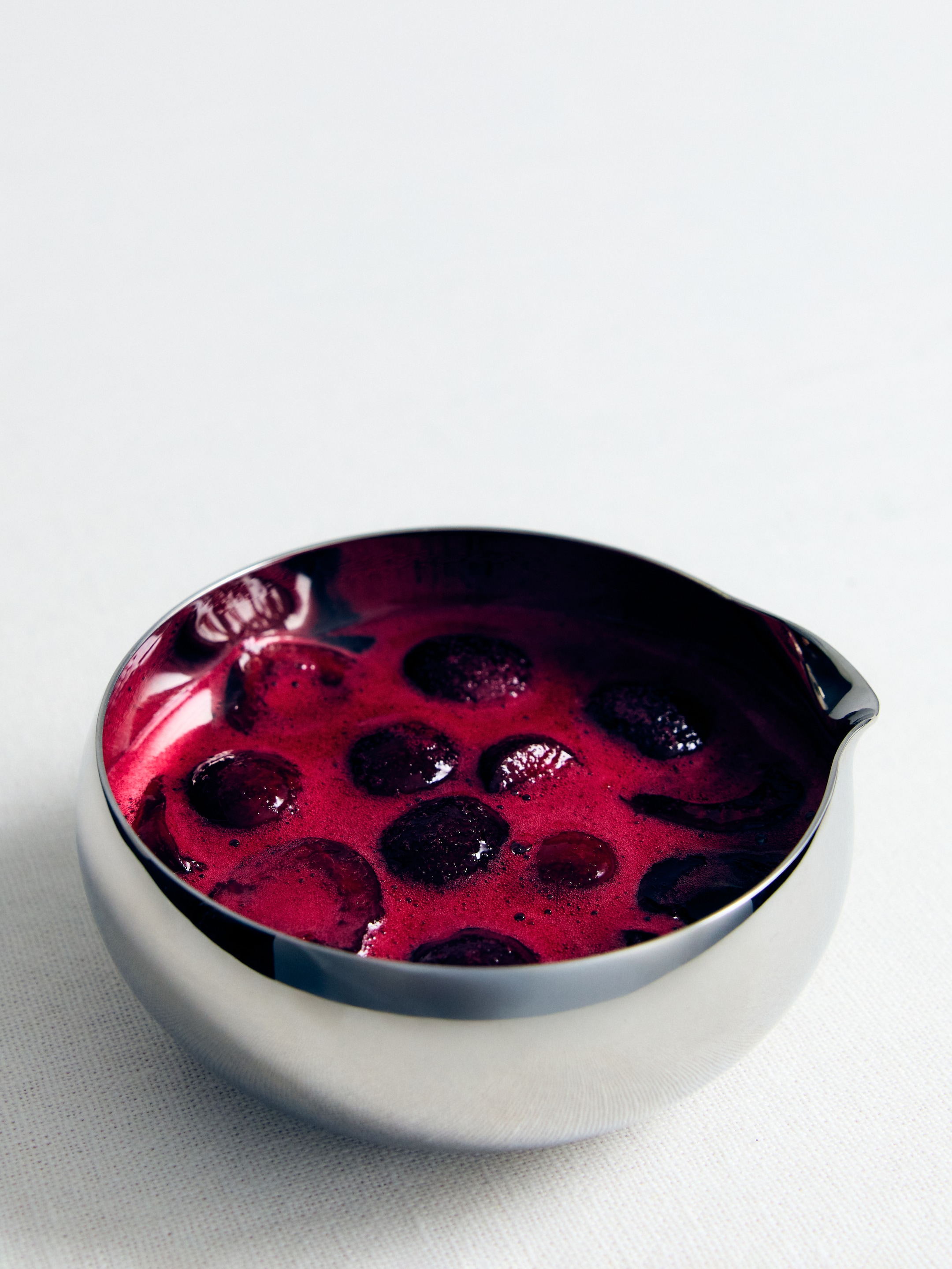 A polished metal bowl filled with a vibrant purple-red sauce, speckled with whole and sliced berries, placed on a white textured surface.
