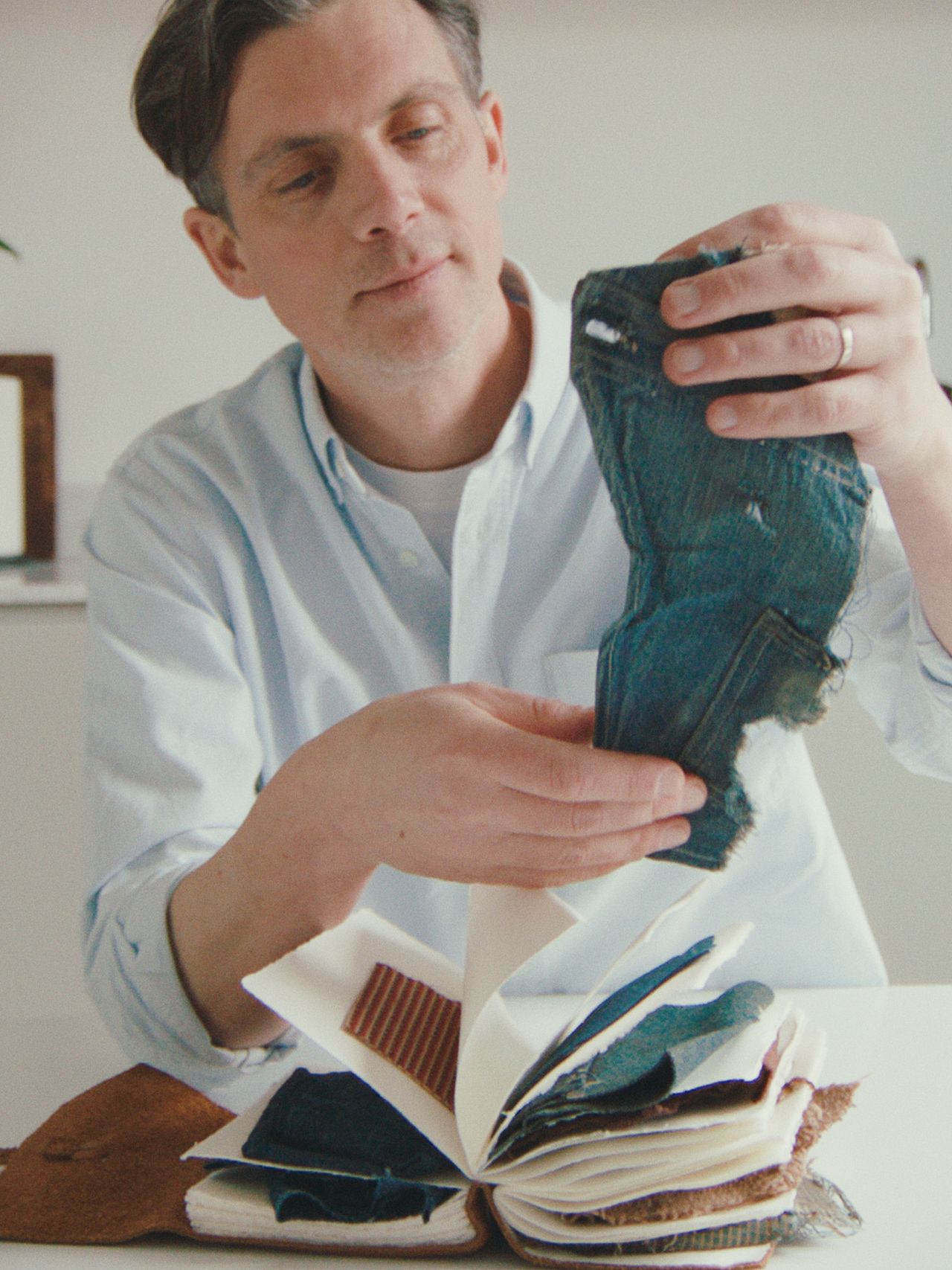 Man examining denim fabric swatch over a book with various fabric samples on a table in a bright room.