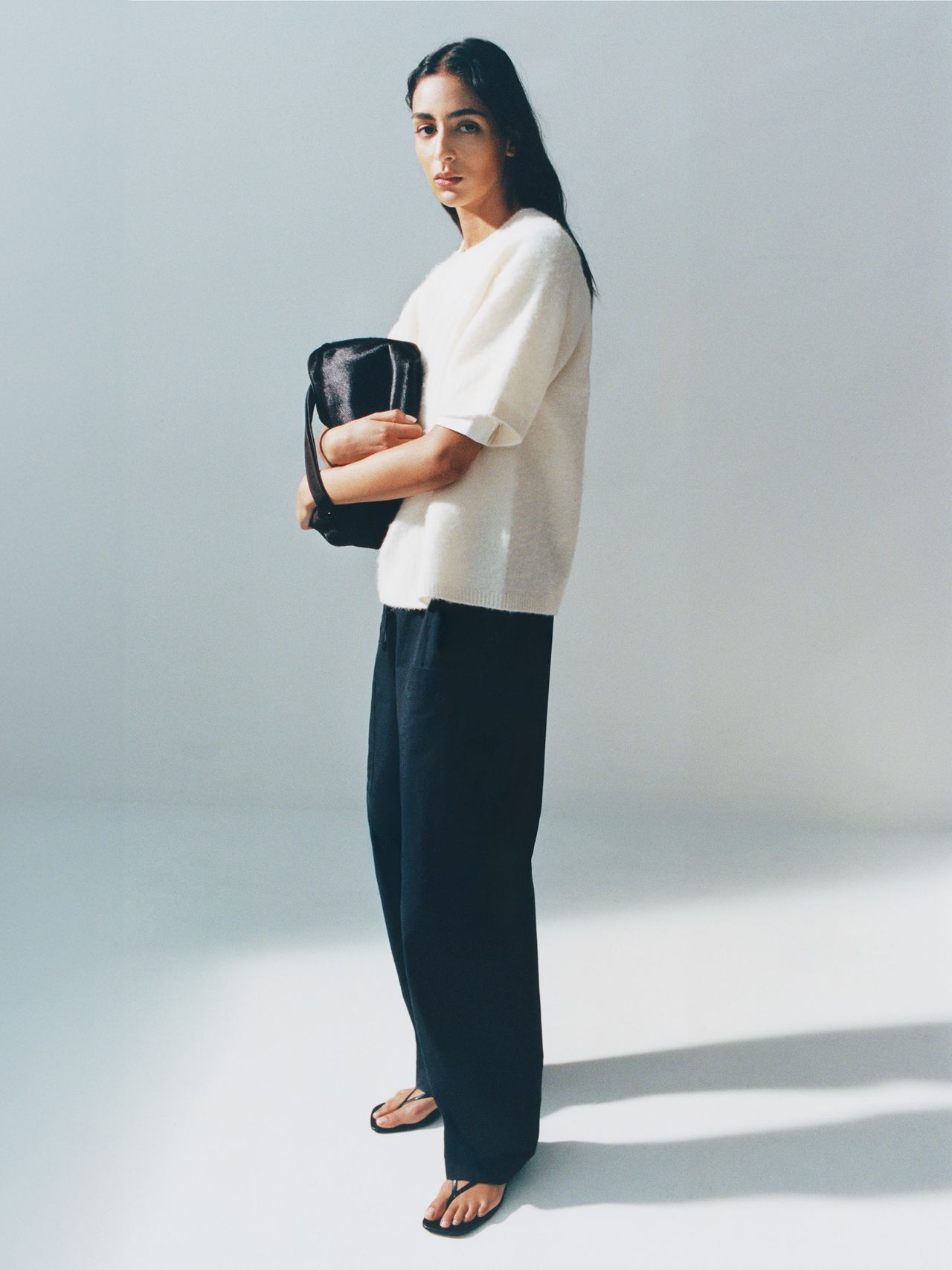 Woman in a white sweater and black pants holds a black bag, standing in a well-lit space with a soft shadow on the floor.