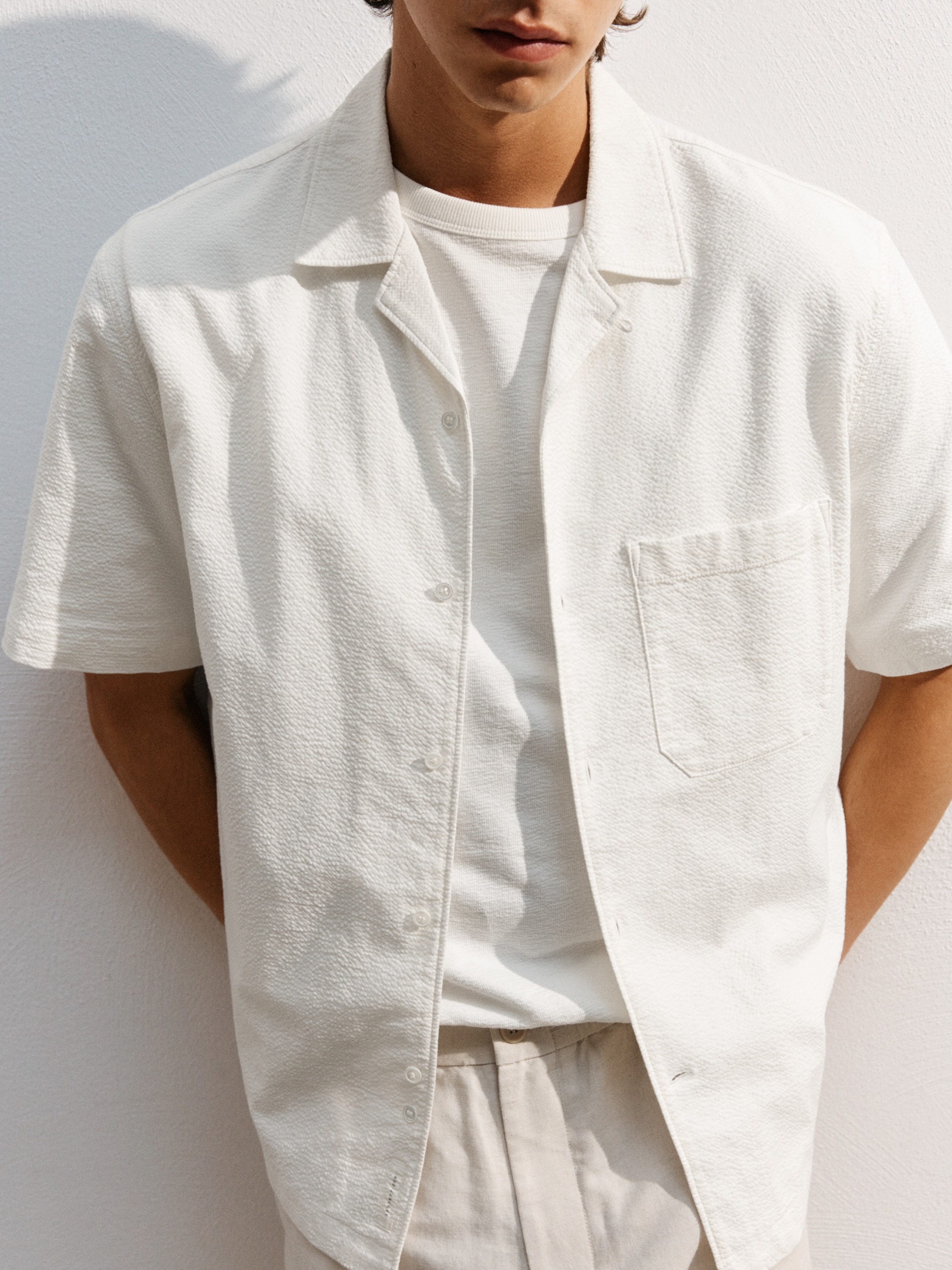 Man in a white short-sleeve shirt over a tee, standing against a white background, hands behind his back, face partially visible.