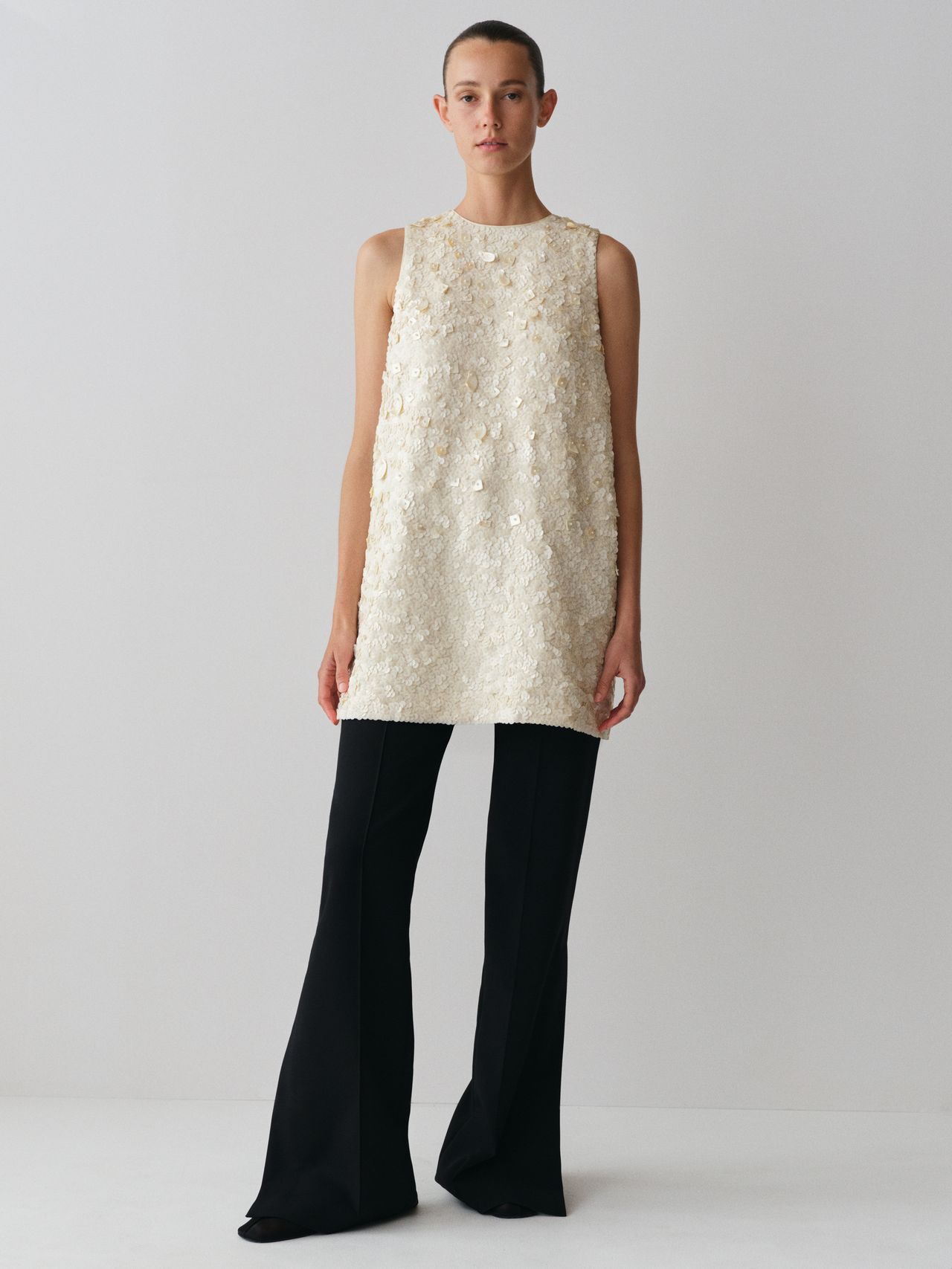 Person wearing a sleeveless, textured cream tunic and black wide-leg pants, standing against a plain white background.