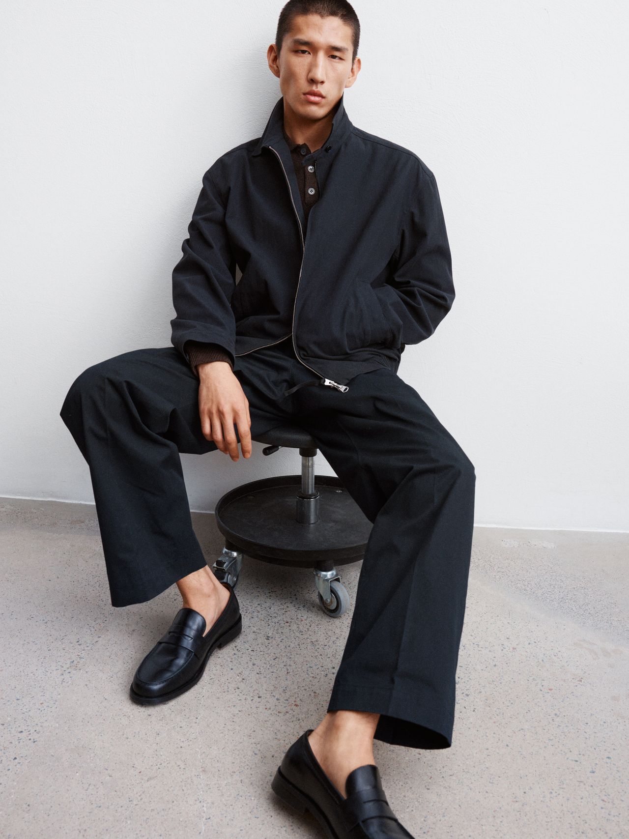 Man in a black outfit sits on a stool, one leg crossed, wearing a jacket, shirt, and loafers, posing against a plain white background.