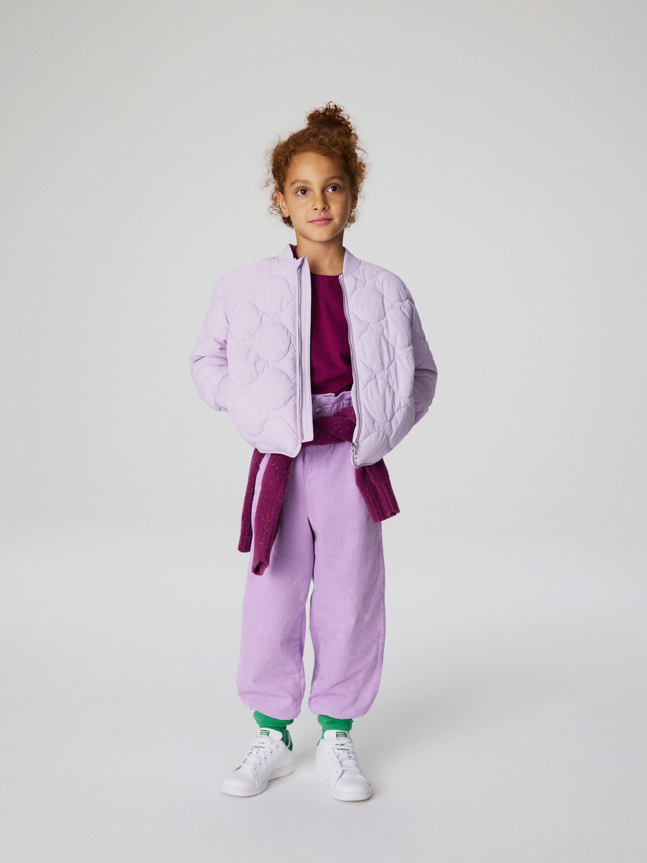 Child in a light purple quilted jacket and pants, maroon shirt, green socks, and white sneakers, standing against a plain background.
