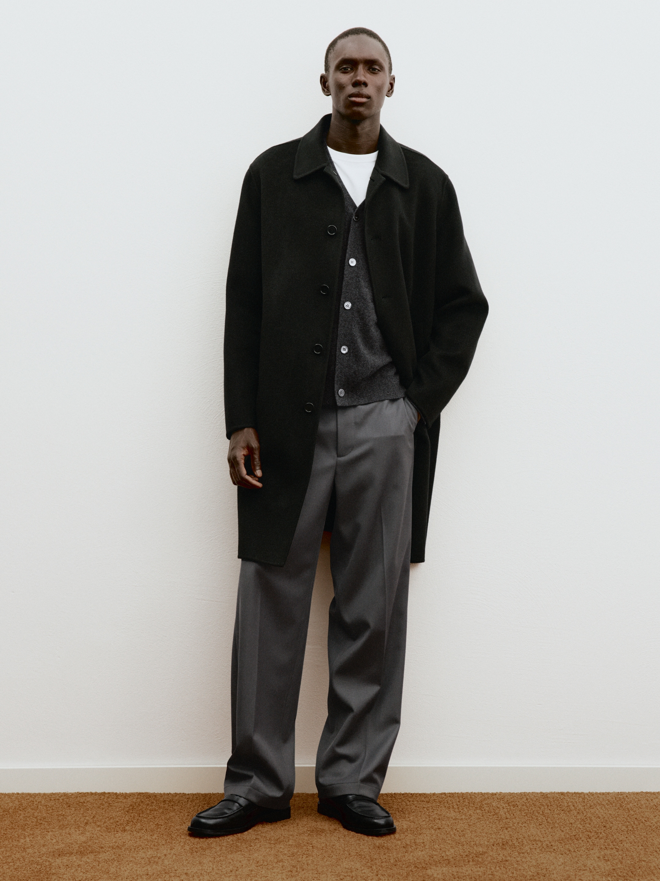 Man in a dark coat and gray trousers stands against a white wall on a brown carpet.