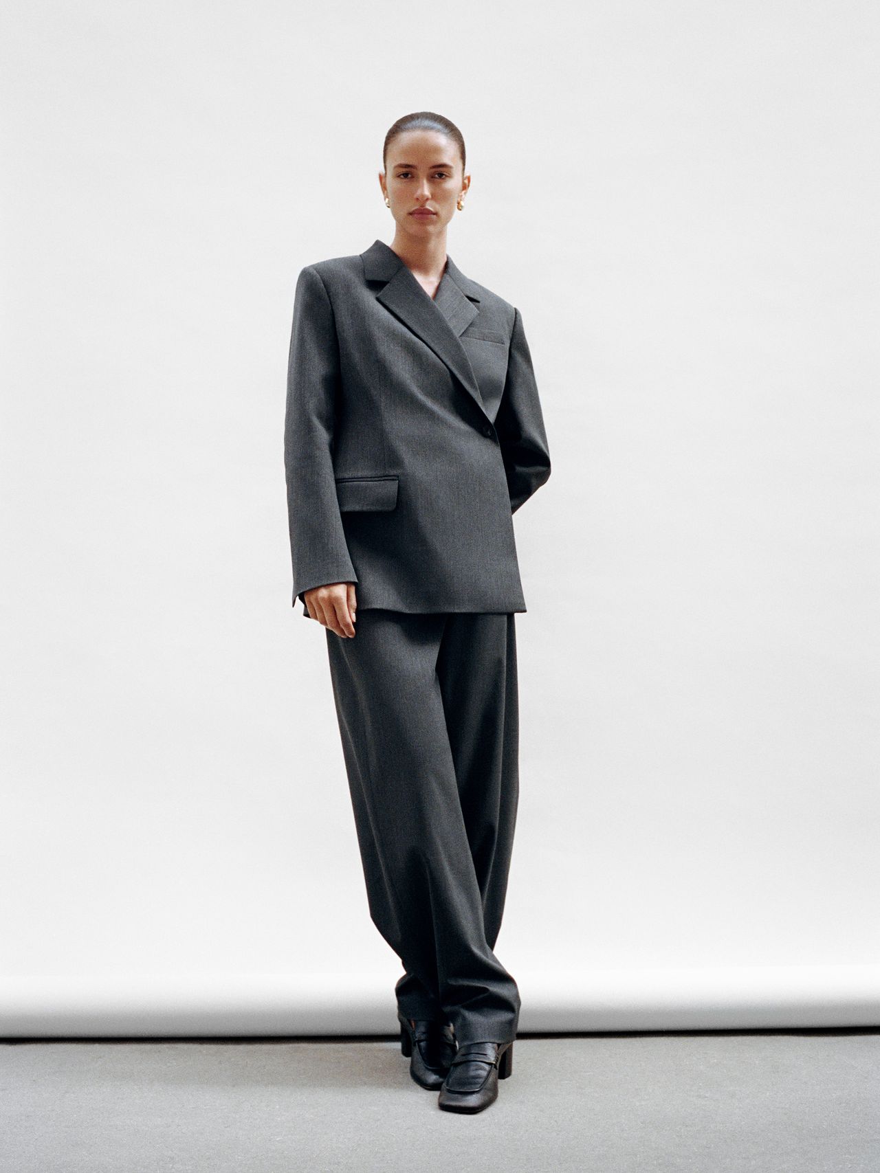 Person in a tailored dark gray suit with wide-legged pants and black shoes, standing against a plain white background.