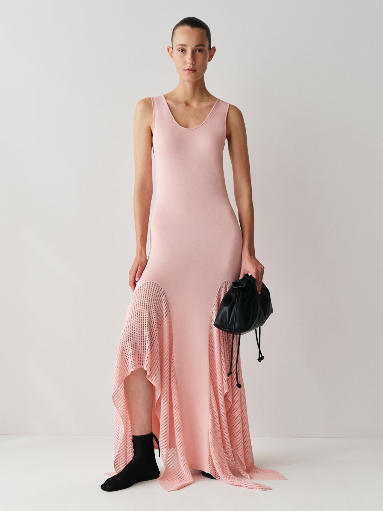 Person in a sleeveless pink dress with a flared hem, holding a black bag, standing against a plain white background.