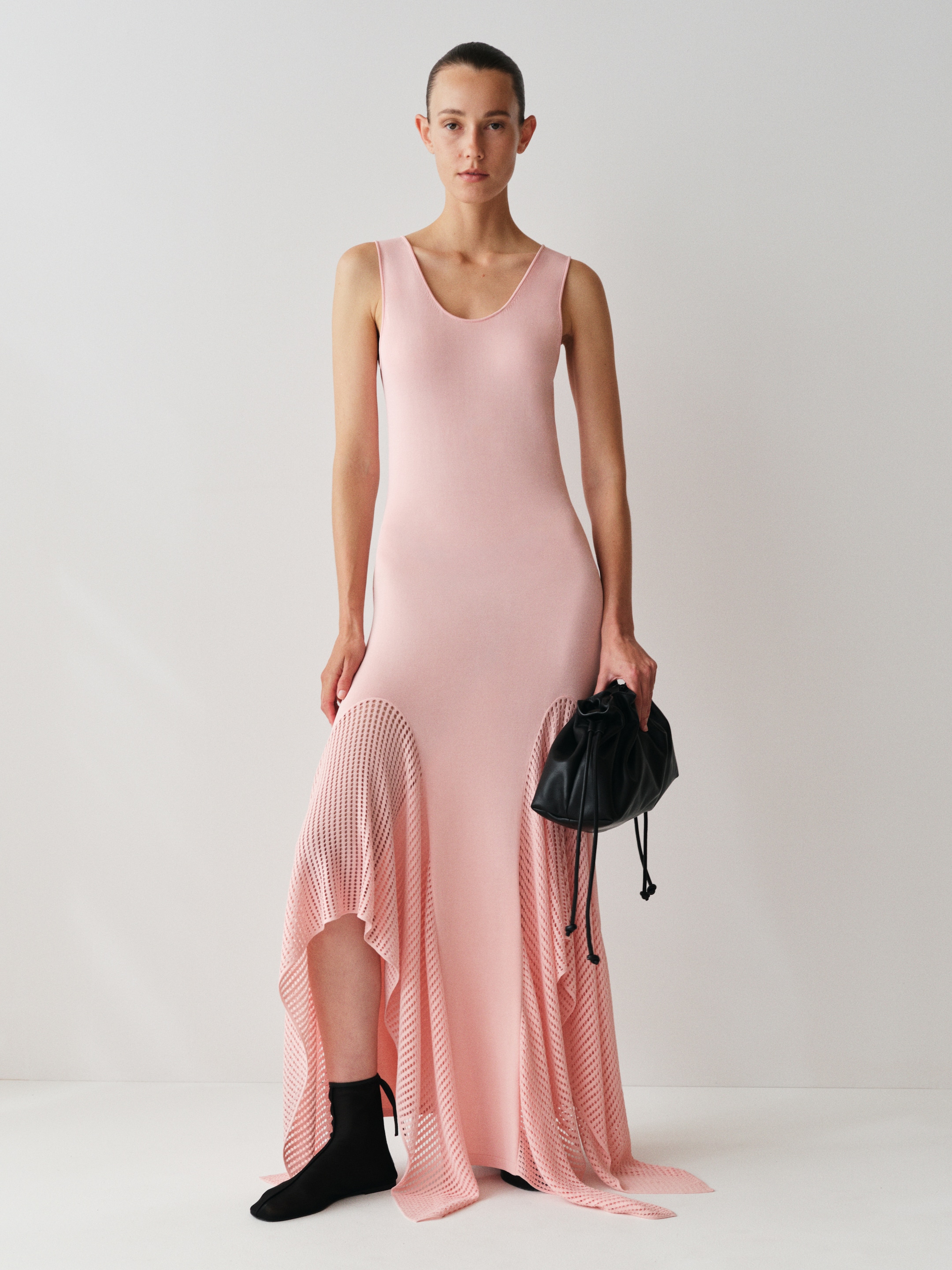 Person in a sleeveless pink dress with a flared hem, holding a black bag, standing against a plain white background.