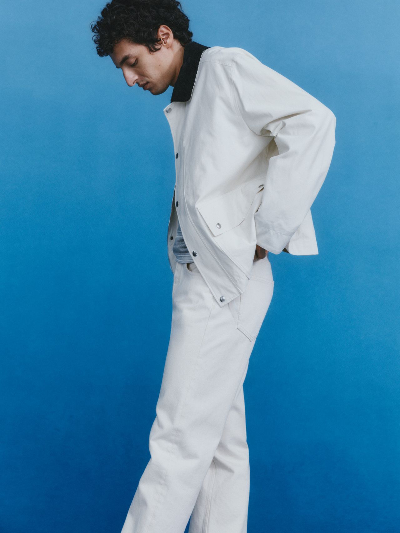Person in a white outfit poses against a blue background, looking down with hands in pockets.