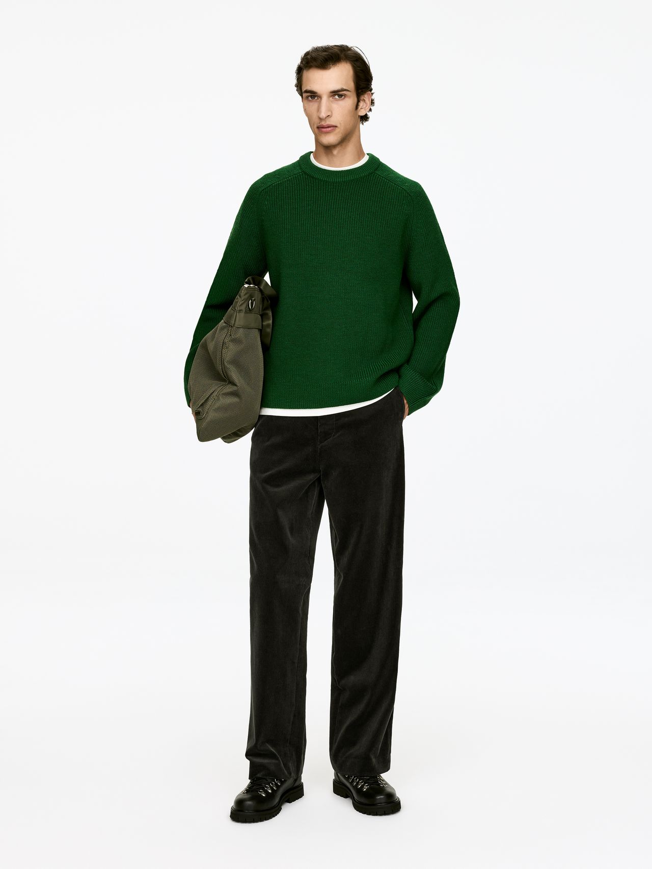 Young man in a green sweater, black corduroy pants and black boots holding an olive duffel bag against a white background.