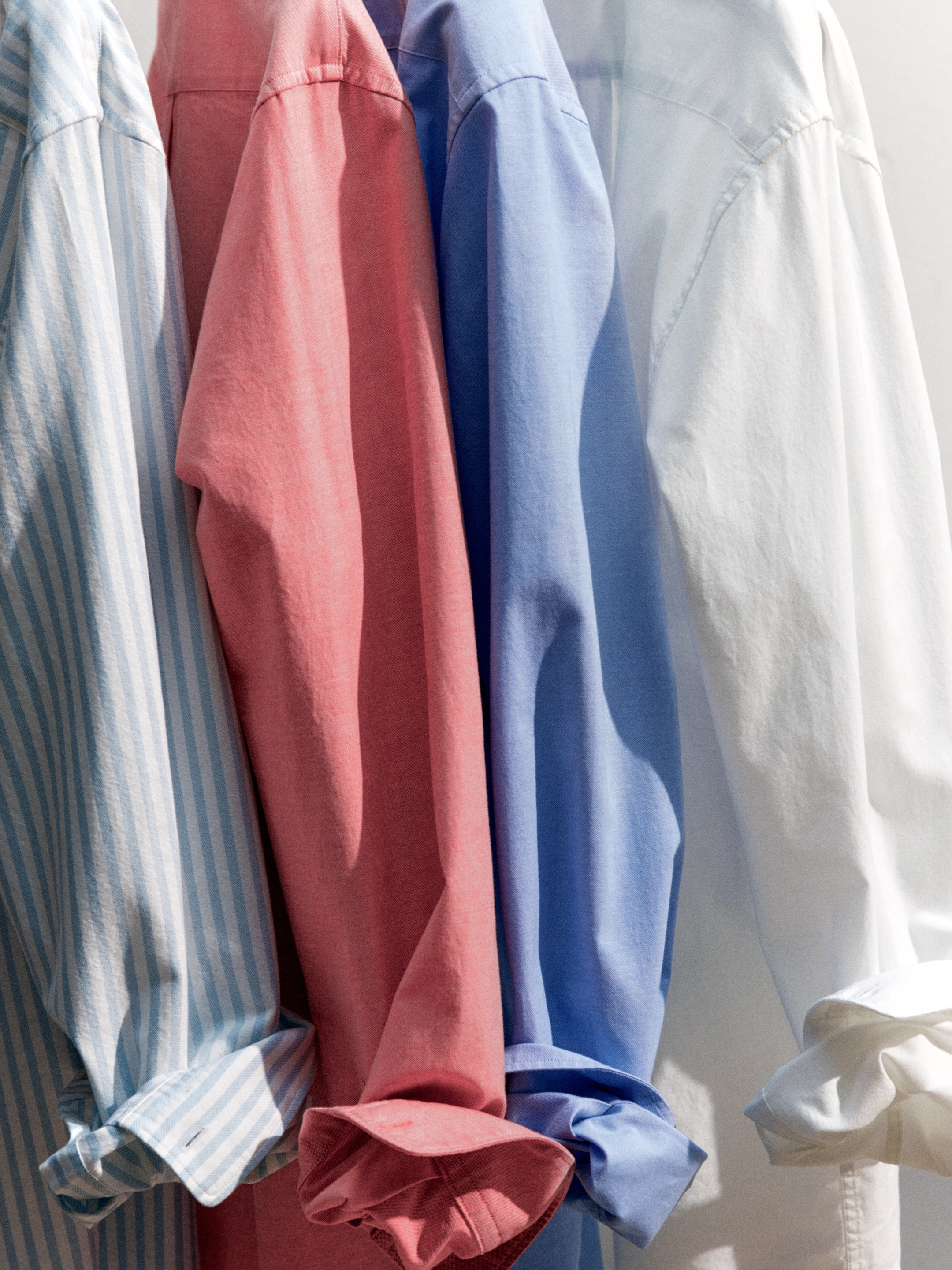 Four shirts hanging side by side: blue striped, pink, blue, and white, with rolled-up sleeves, illuminated by soft light.