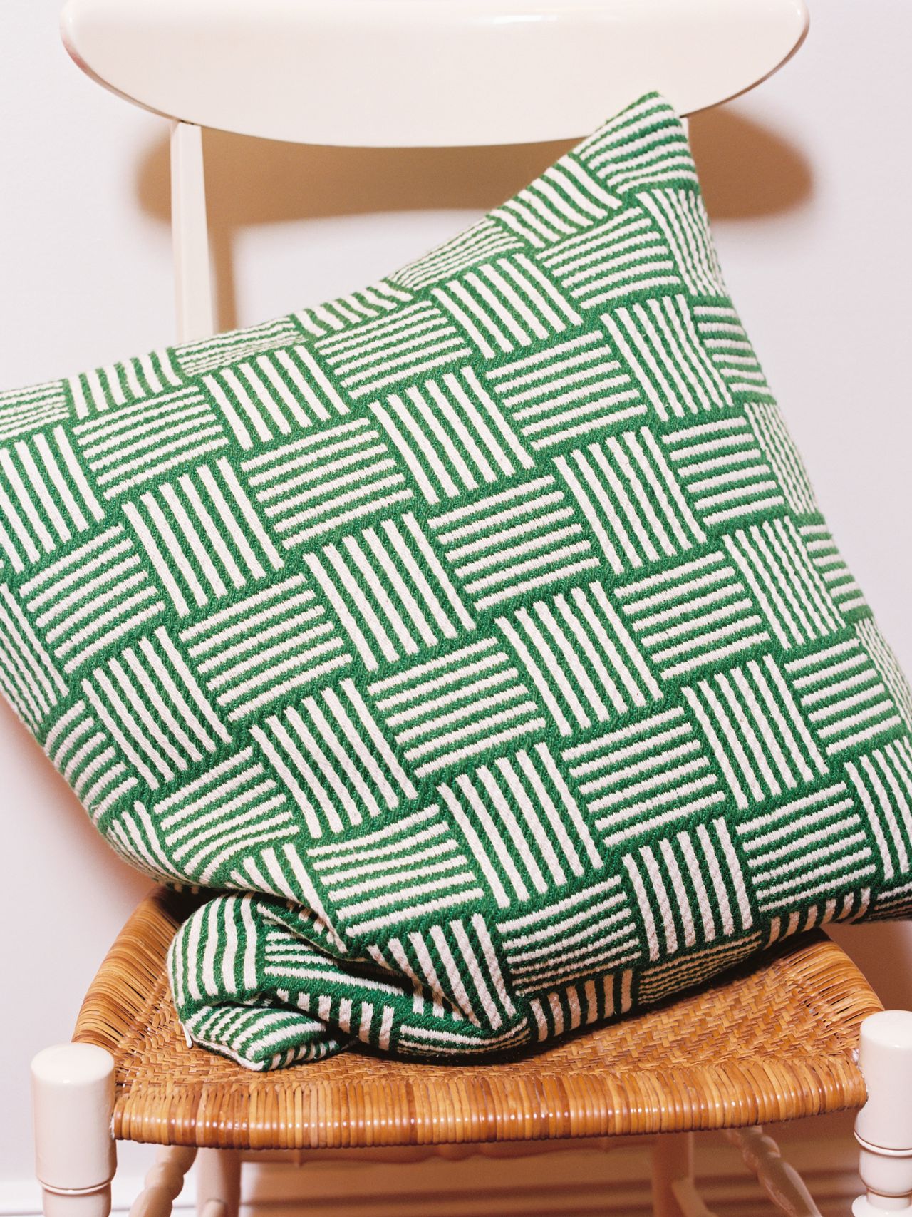Green and white patterned pillow on a wicker chair with a white backrest, against a plain white wall.