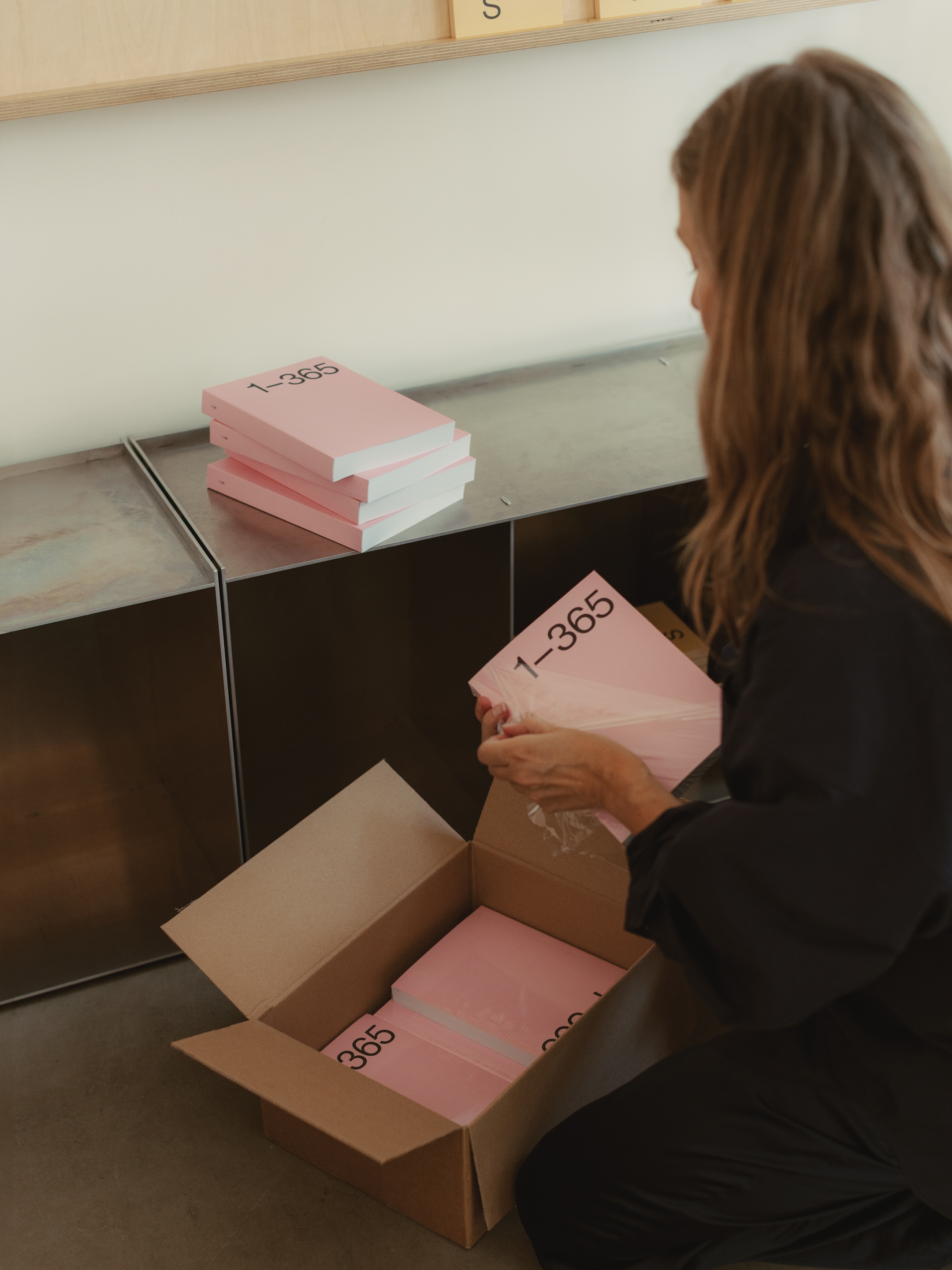 A woman kneels beside an open cardboard box, unpacking pink books labeled "1-365," with more books stacked on a metal shelf nearby.