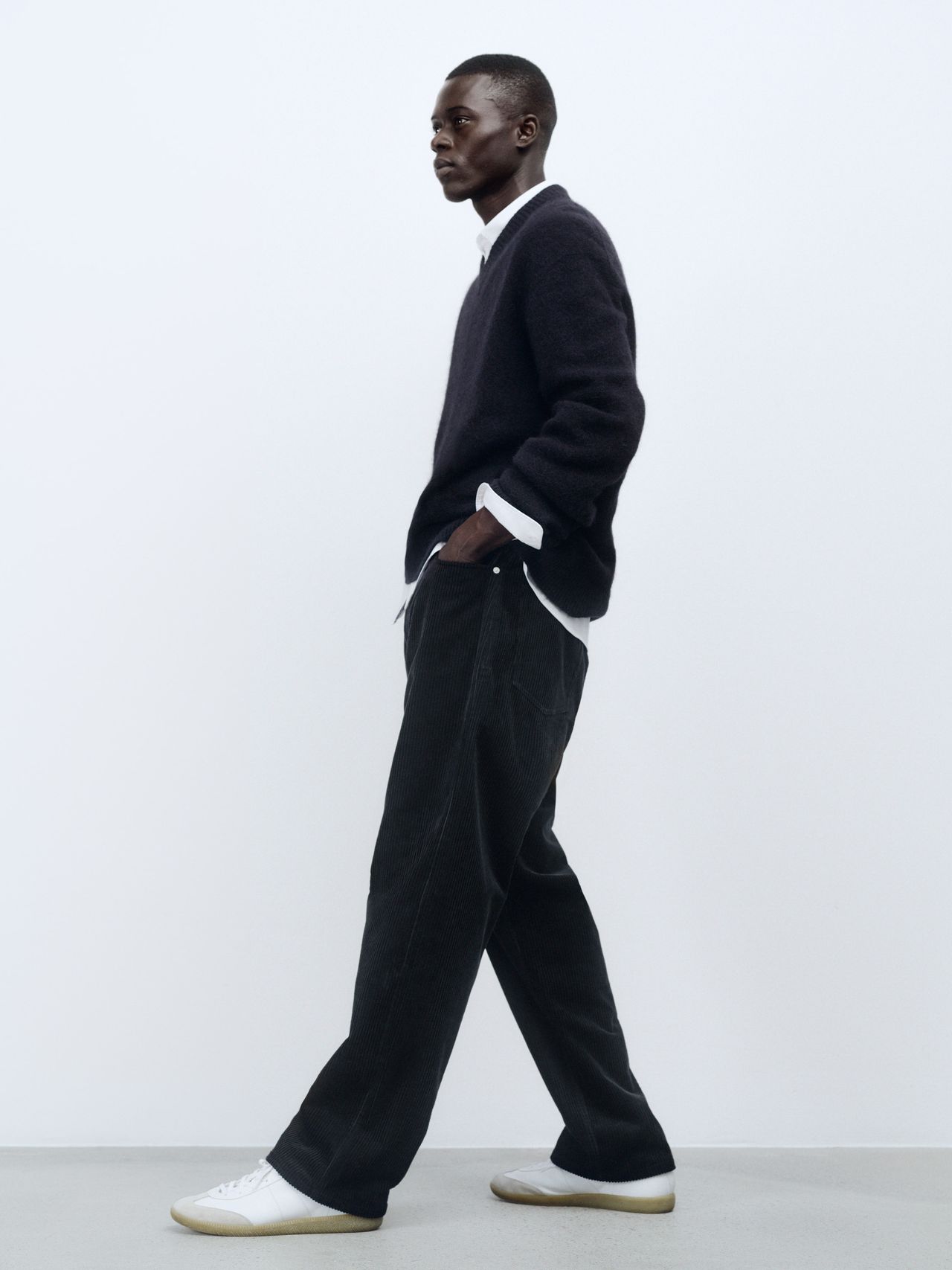 Man in a dark sweater and wide-leg pants, walking sideways against a plain white background, hands in pockets, wearing white sneakers.