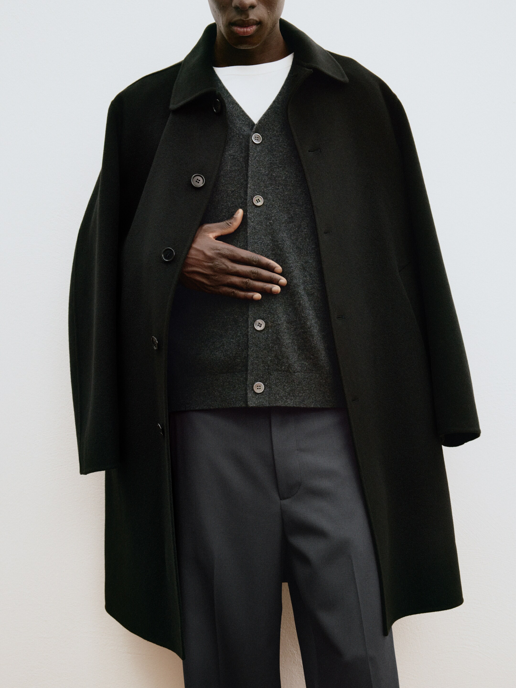 A person dressed in a dark overcoat, gray cardigan, and trousers, standing against a neutral background. Their hand rests on their midsection.