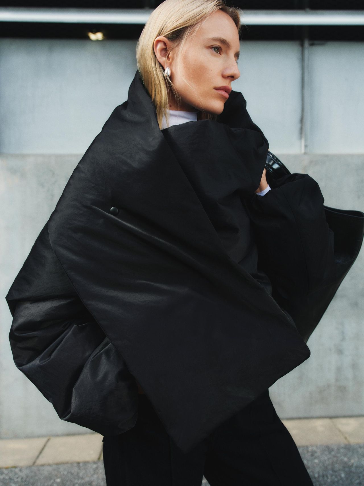 Person in a large, black, puffy coat stands outdoors, looking to the side, with a modern building backdrop.