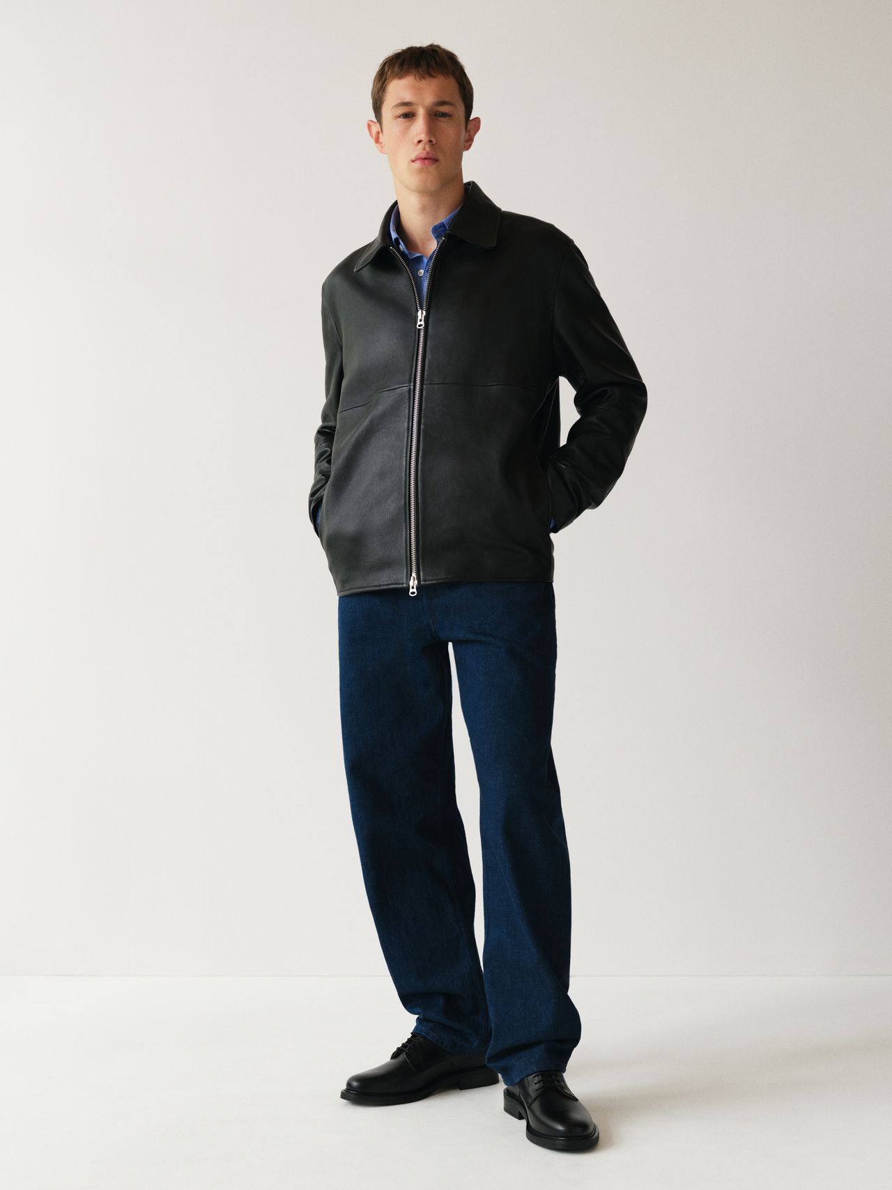 A person stands against a plain background, wearing a black leather jacket, blue jeans, and black shoes, with hands in pockets.