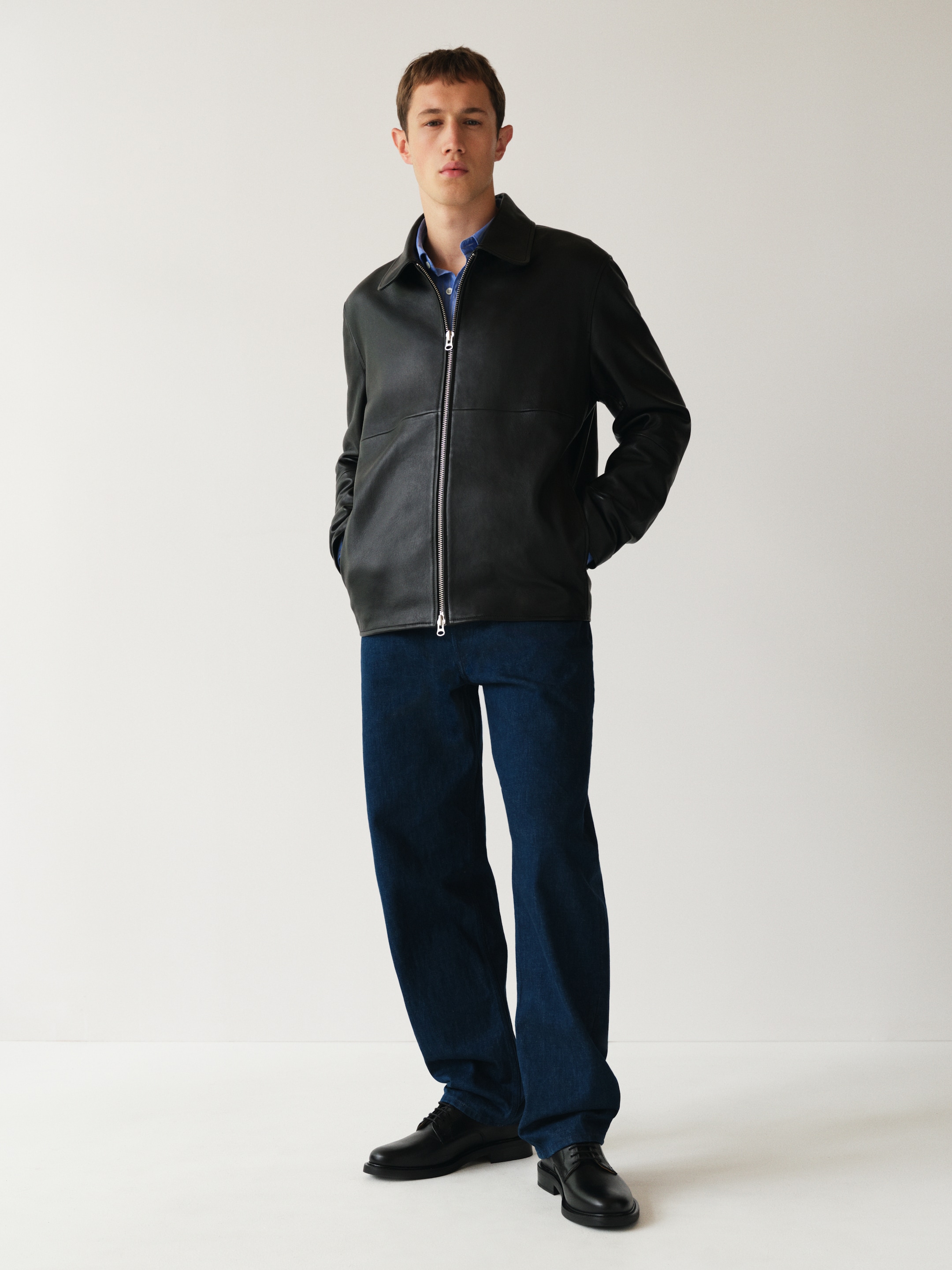 A person stands against a plain background, wearing a black leather jacket, blue jeans, and black shoes, with hands in pockets.