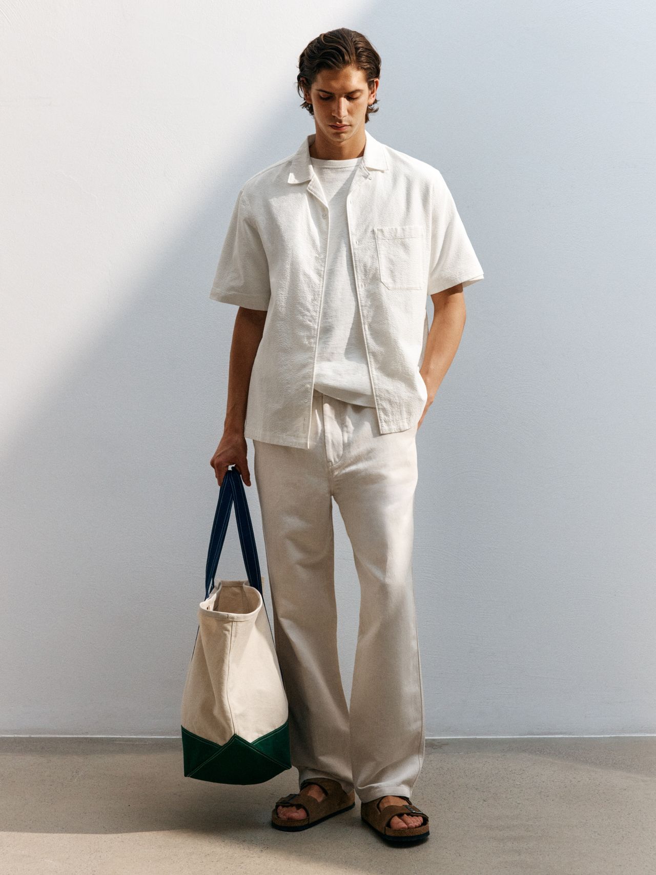 A person wearing a white shirt and pants, holding a canvas tote bag with blue and green accents, stands against a light wall.
