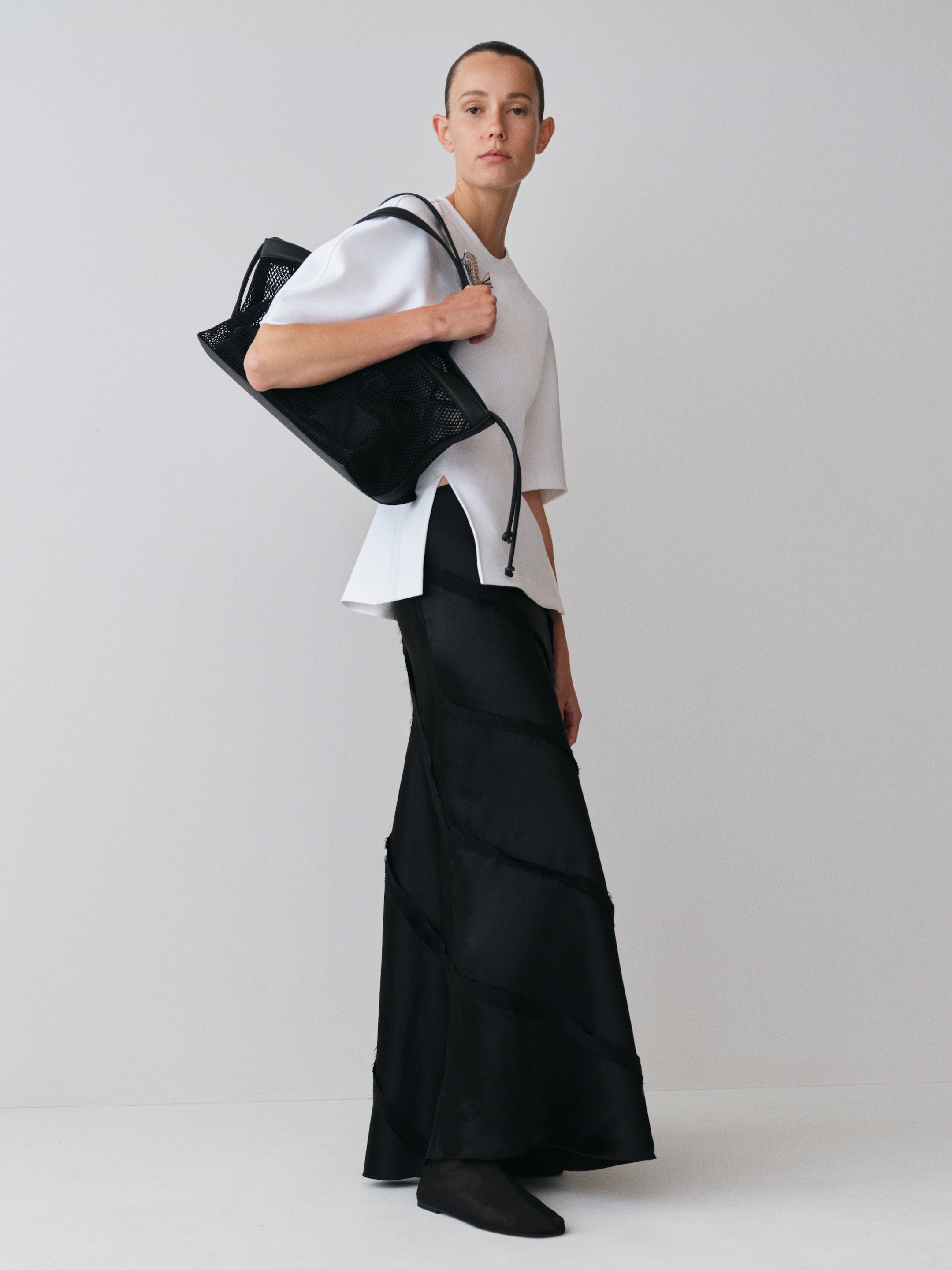 Person in a white top and long black skirt, holding a black bag, stands against a plain white background.