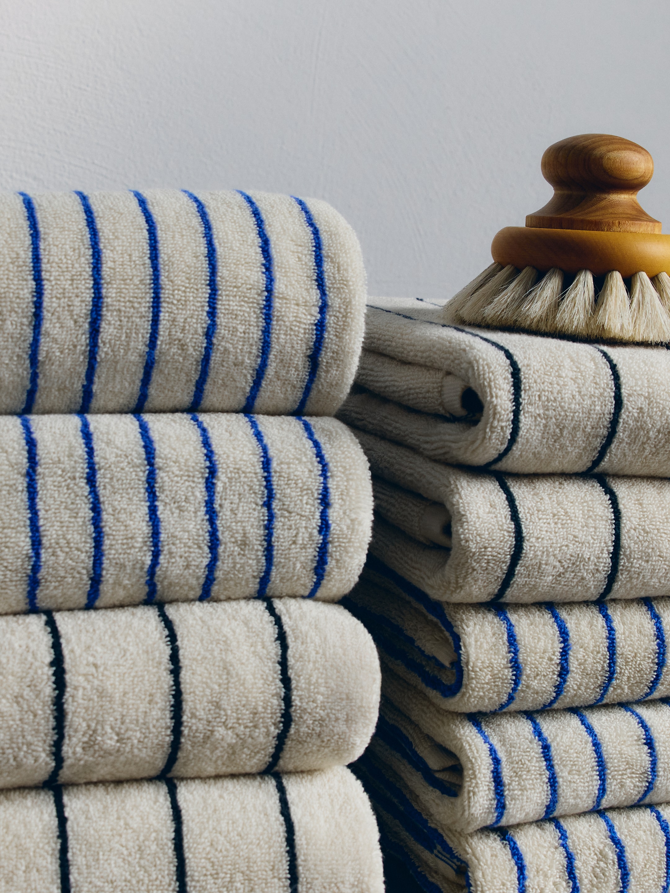 Stacked cream towels with blue and black stripes beside a brush with wooden handle, set against a plain white wall.