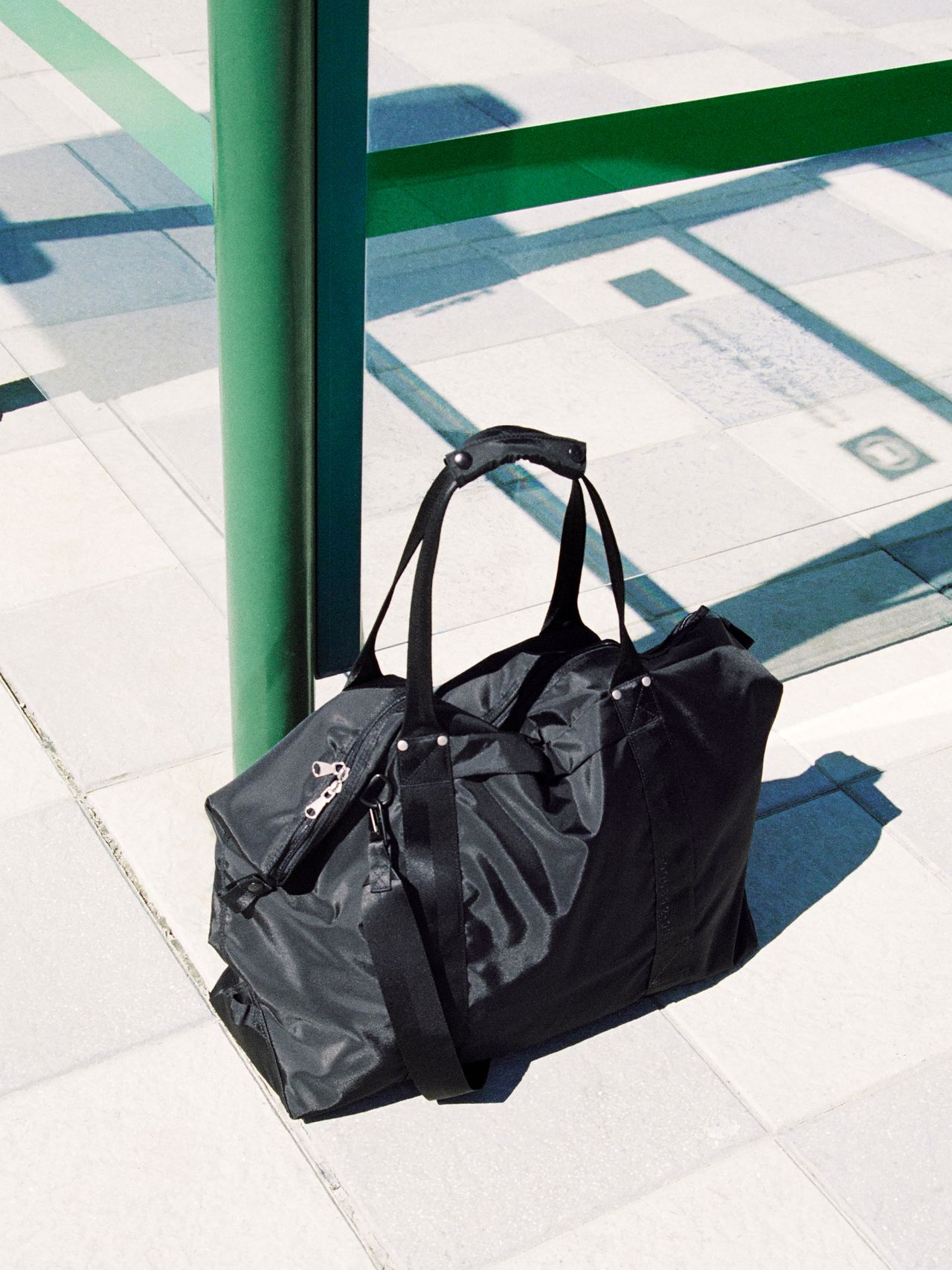 Black duffel bag with handles and a shoulder strap, placed on a sunlit tiled pavement near a green pole.