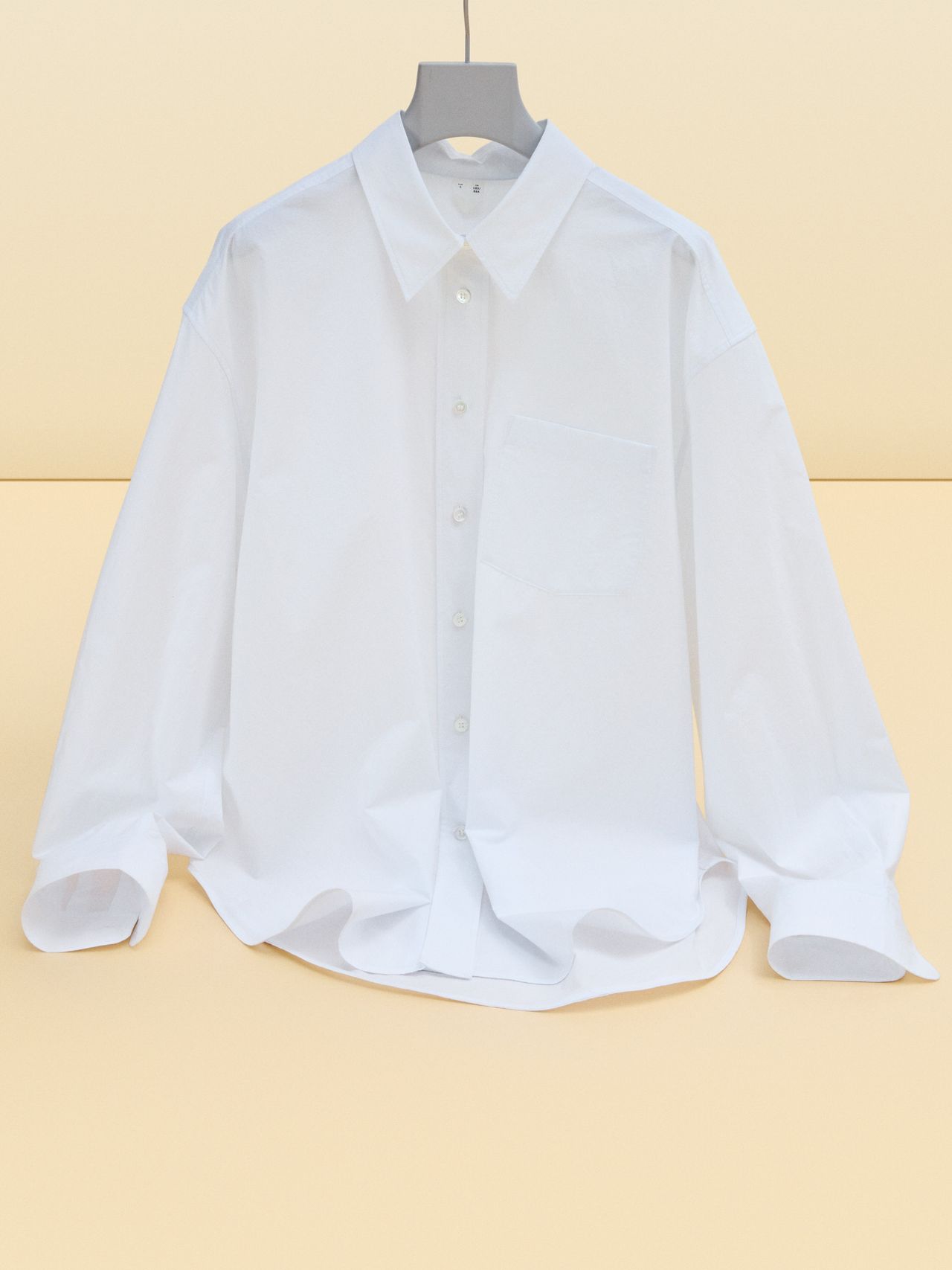 A crisp white button-up shirt on a hanger, with long sleeves and a collar, against a soft beige background.