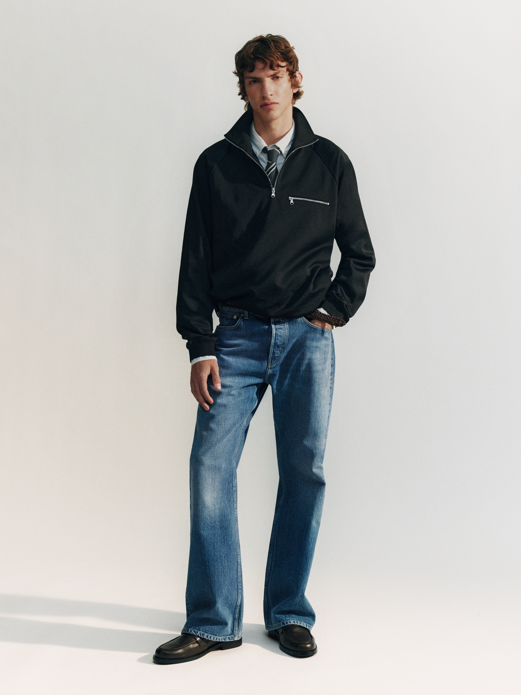 A person wearing a black pullover, light shirt, tie, blue jeans, and black shoes stands against a plain backdrop.
