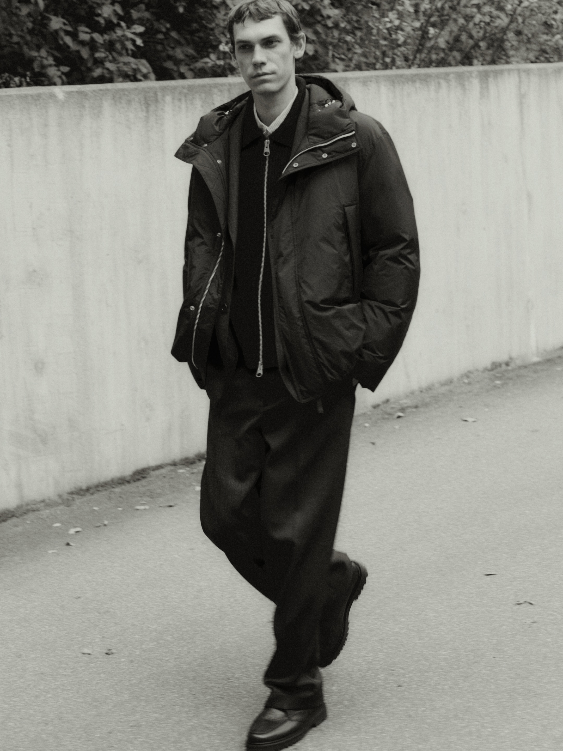 A person in a dark jacket and pants walks casually beside a concrete wall with foliage in the background. The image is in black and white.