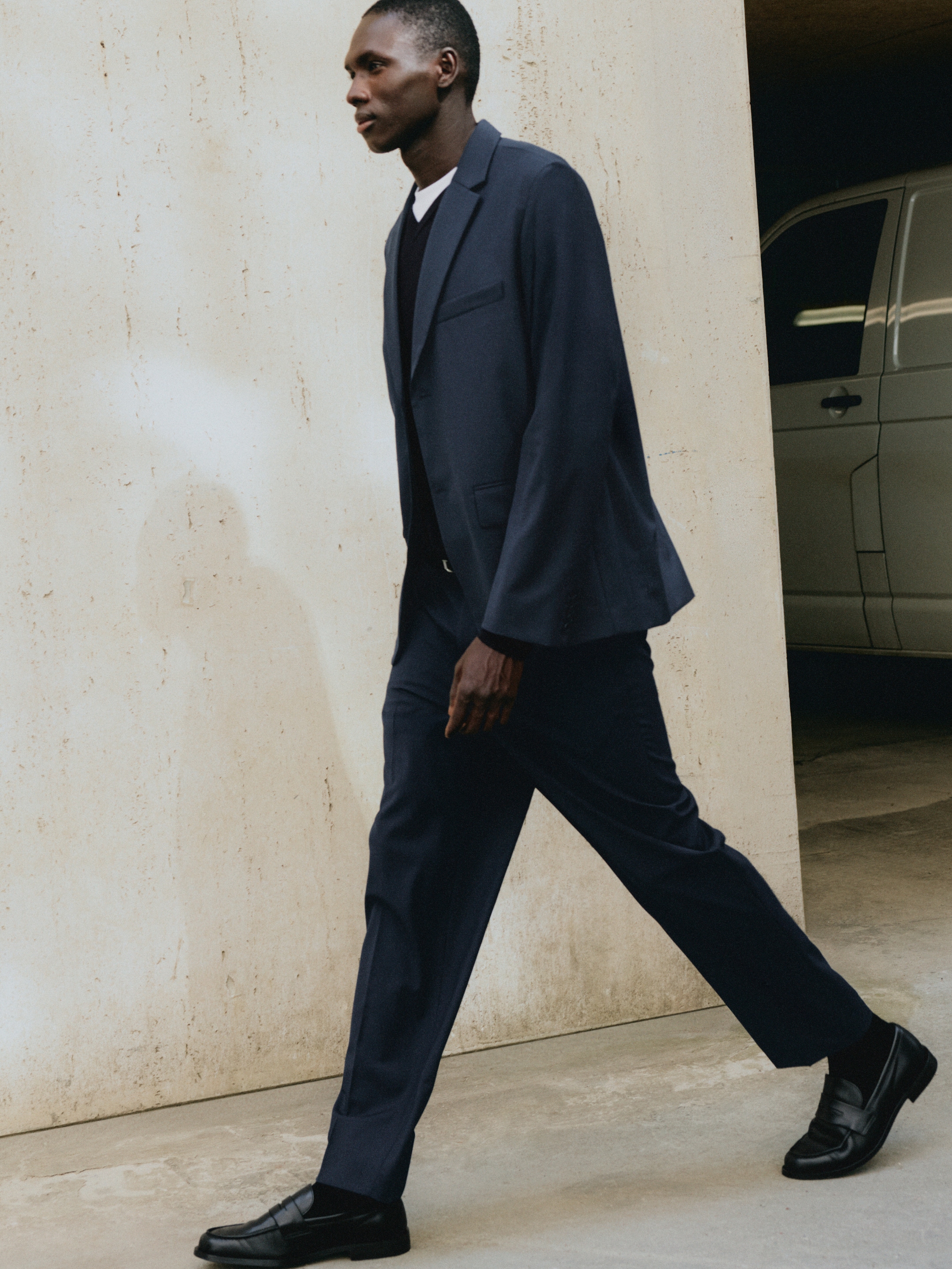 A person walking confidently in a dark suit and loafers past a concrete wall, with part of a white van visible in the background.