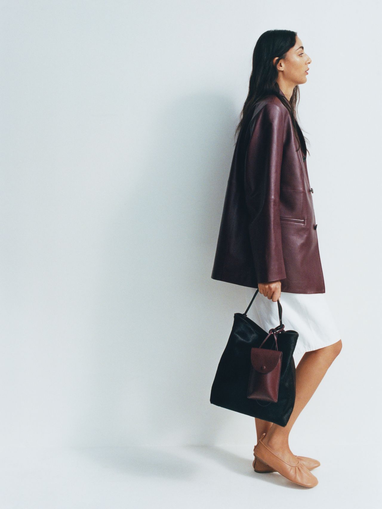 A person in a burgundy leather jacket and white skirt holds a black and burgundy tote bag, standing against a plain white background.