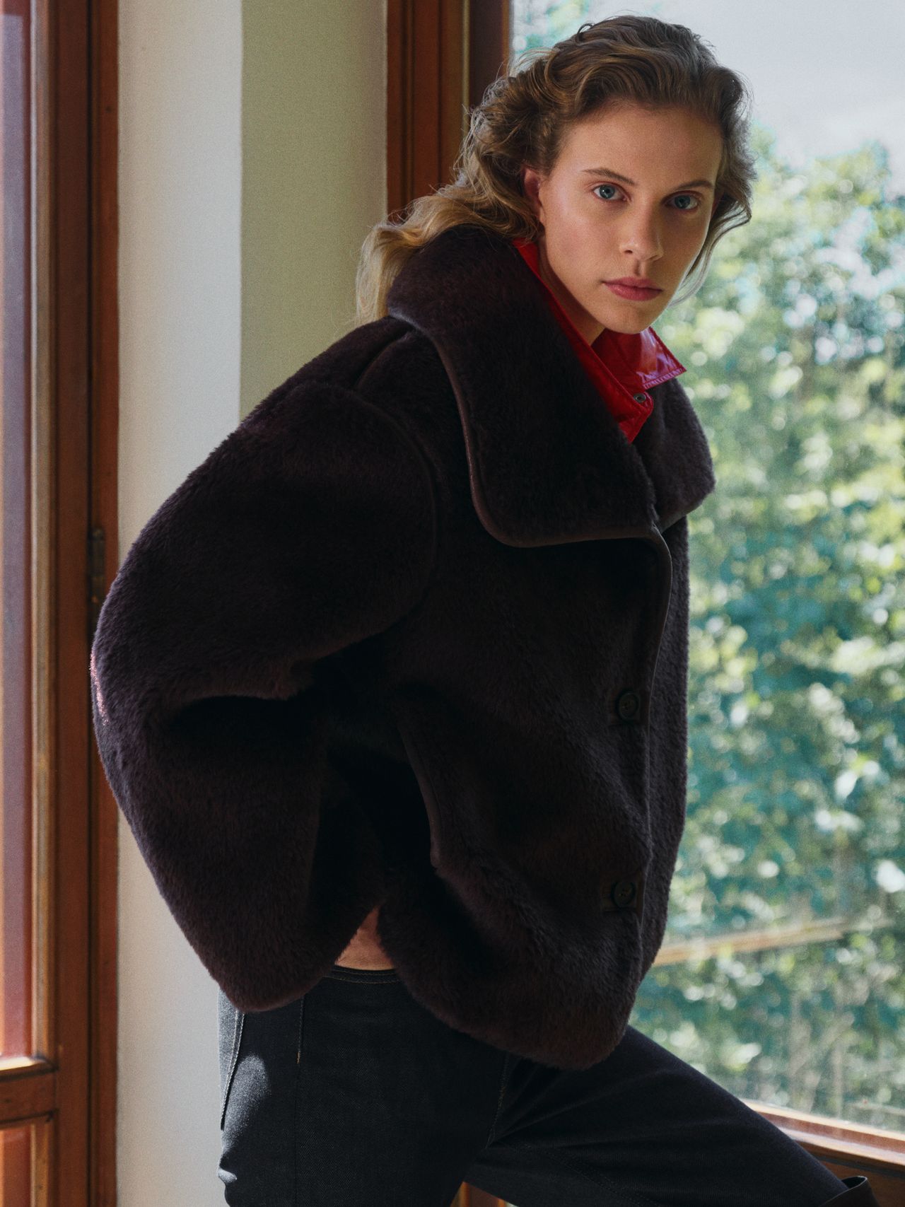 Person wearing a dark fur coat over a red shirt, standing by a window with trees visible outside.