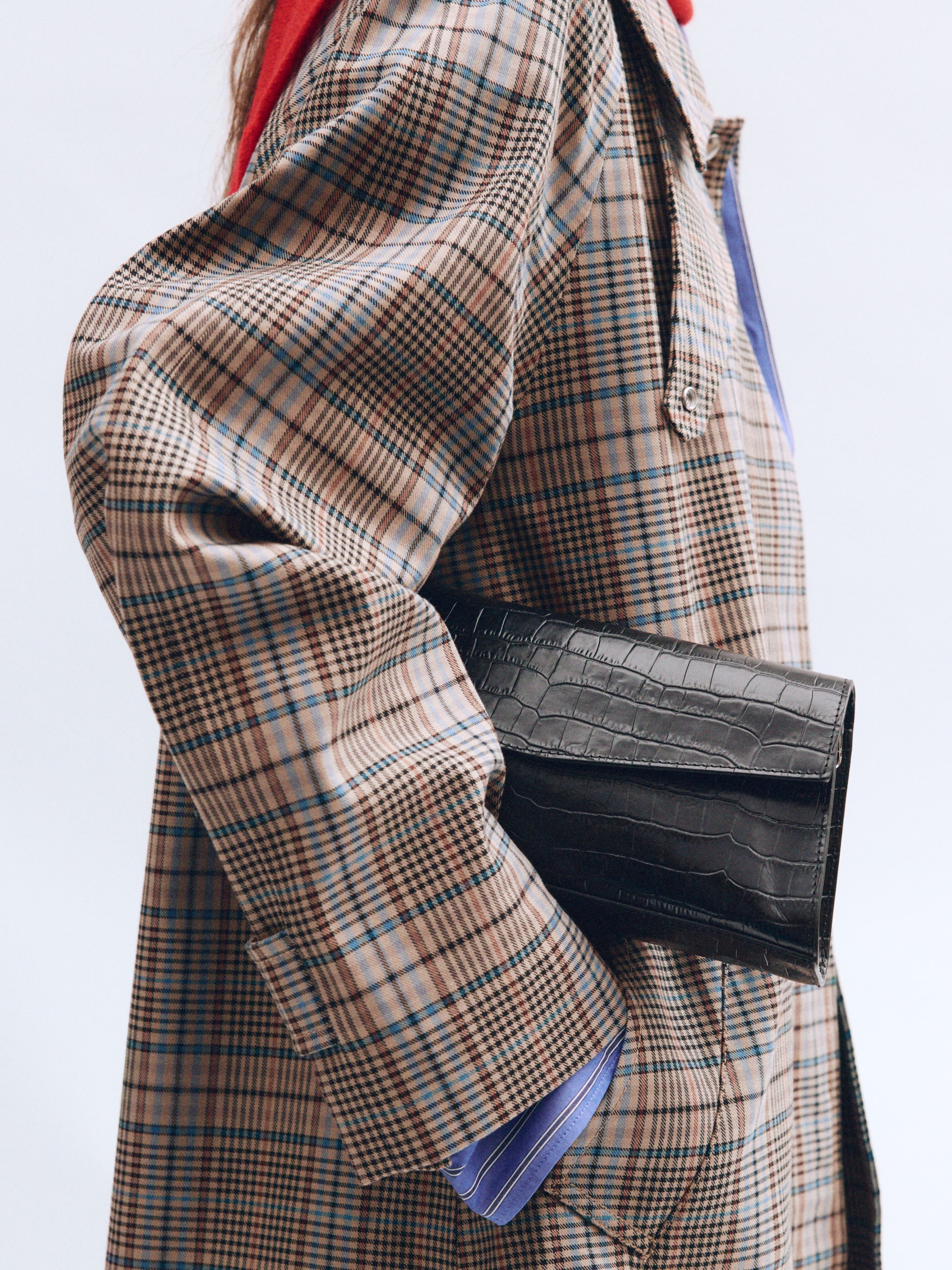 Person wearing a plaid coat with a black crocodile-patterned clutch tucked under their arm, against a light background.