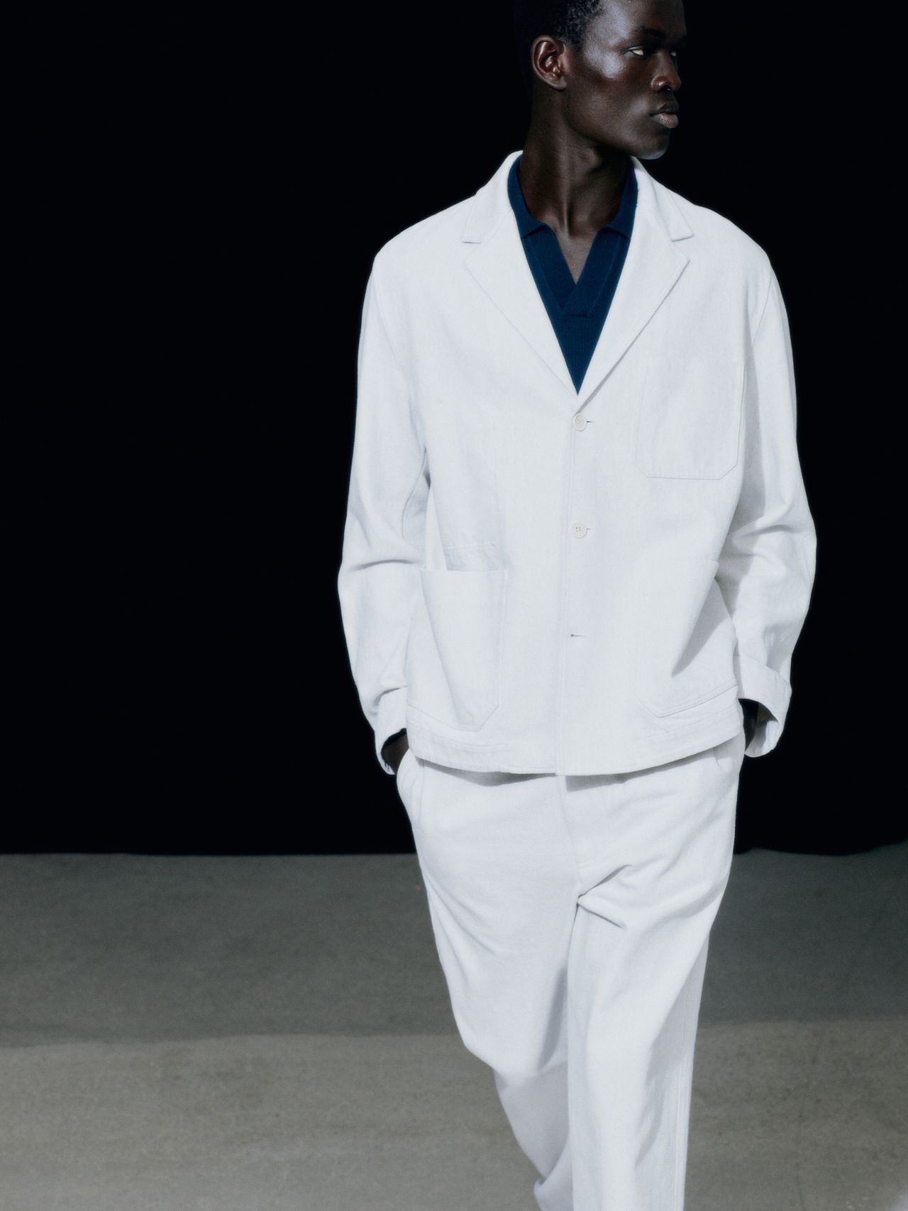 Person in white suit with blue shirt underneath, standing against a dark background, looking to the side.