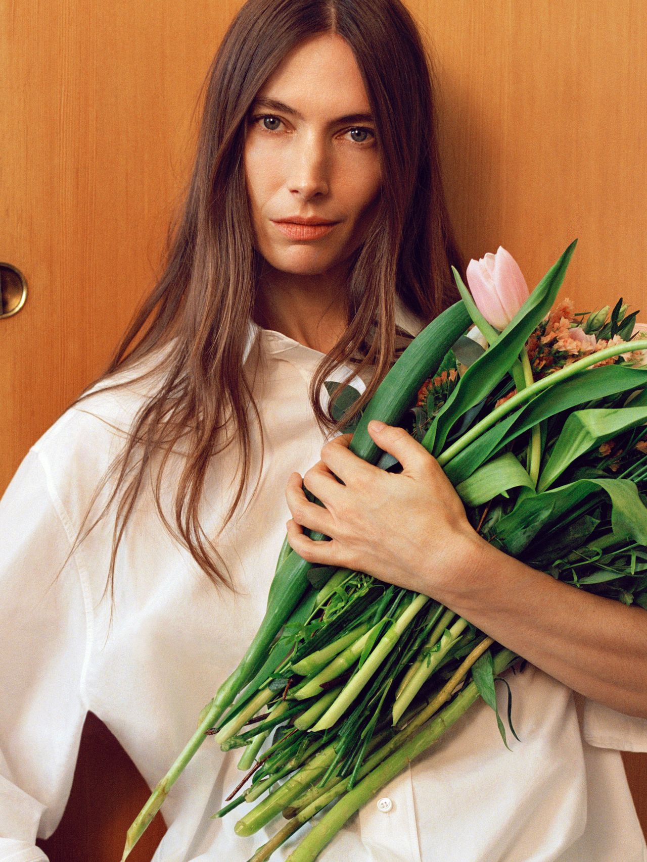 Woman with long hair holding a bouquet of green leaves and a pink tulip, wearing a white shirt, standing against a wooden background.