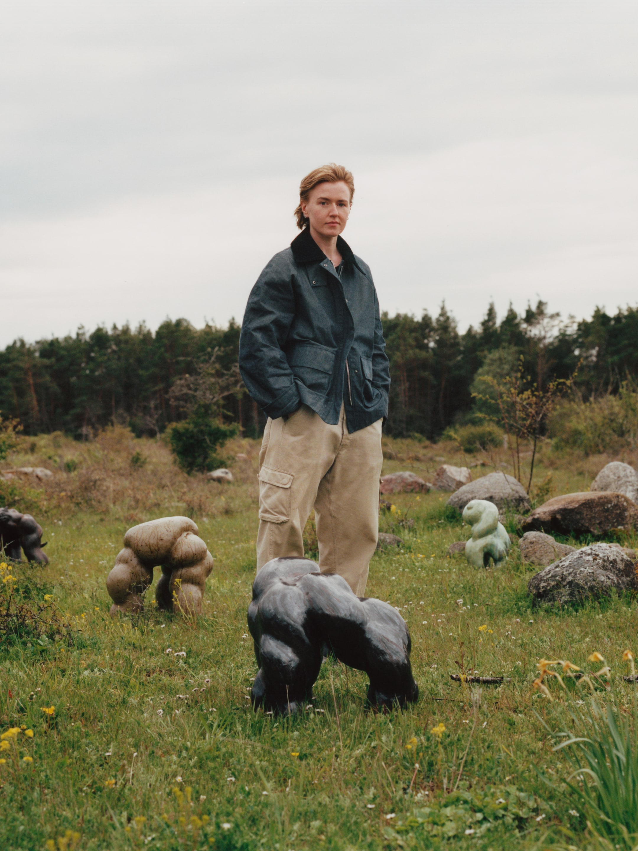 Person in a field with small, abstract animal sculptures, wearing a dark jacket and beige pants. Trees and cloudy sky in the background.