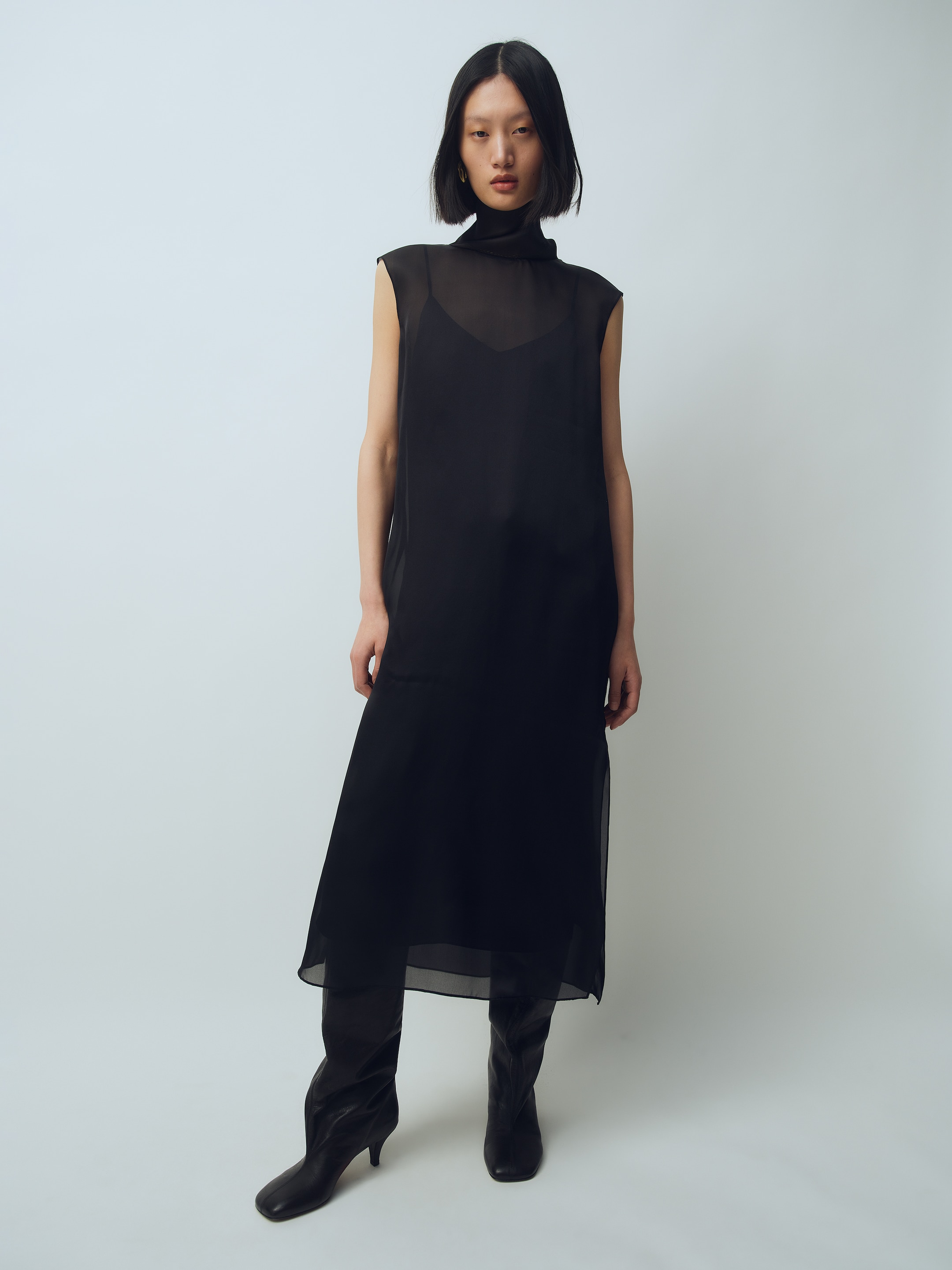 A woman with short black hair wears a sleeveless, sheer black dress over a dark outfit, paired with knee-high black boots, against a light background.