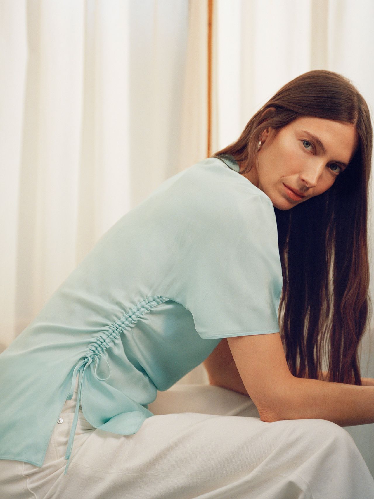 A person with long brown hair in a light blue ruched top and white pants, sitting in a softly lit room with cream-colored curtains.