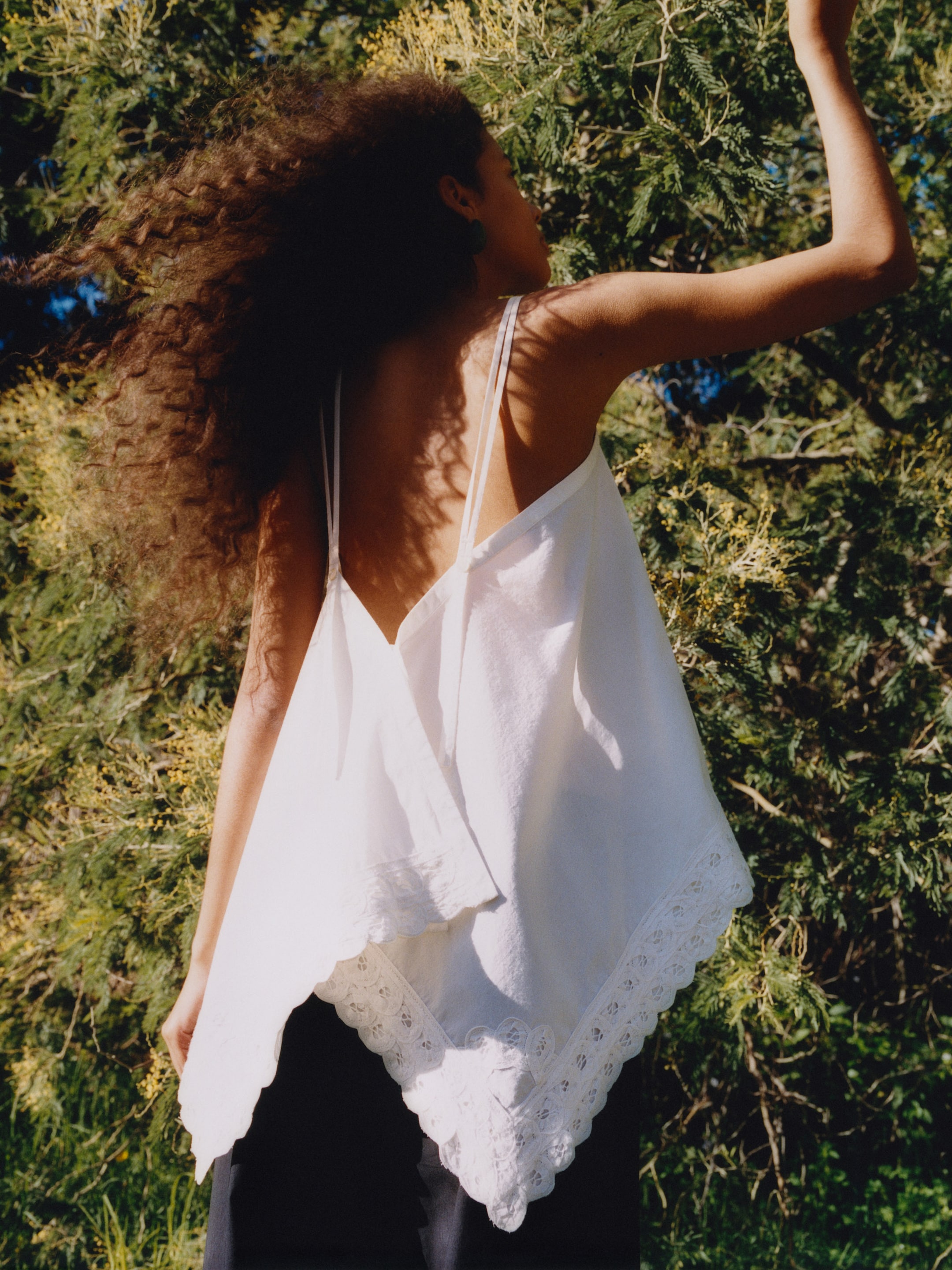 Woman with curly hair wearing a white, flowy top stands outdoors, surrounded by greenery, facing away from the camera.