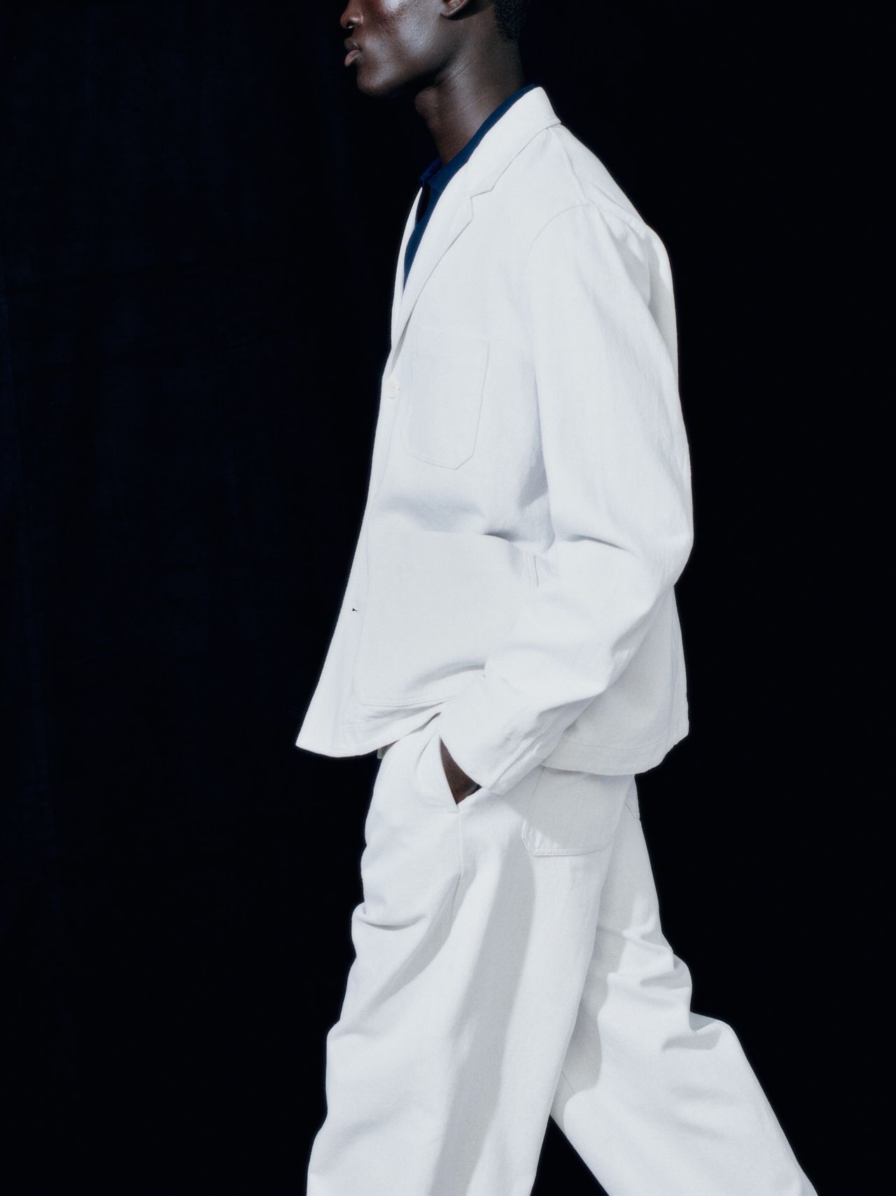 Person in a white suit and dark shirt against a black background, walking with hands in pockets, profile view.