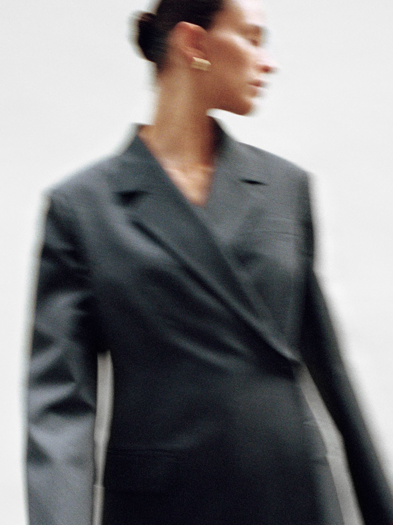 A person in a blurred image wears a dark, oversized blazer. The background is plain white, and the person's face is turned to the side.