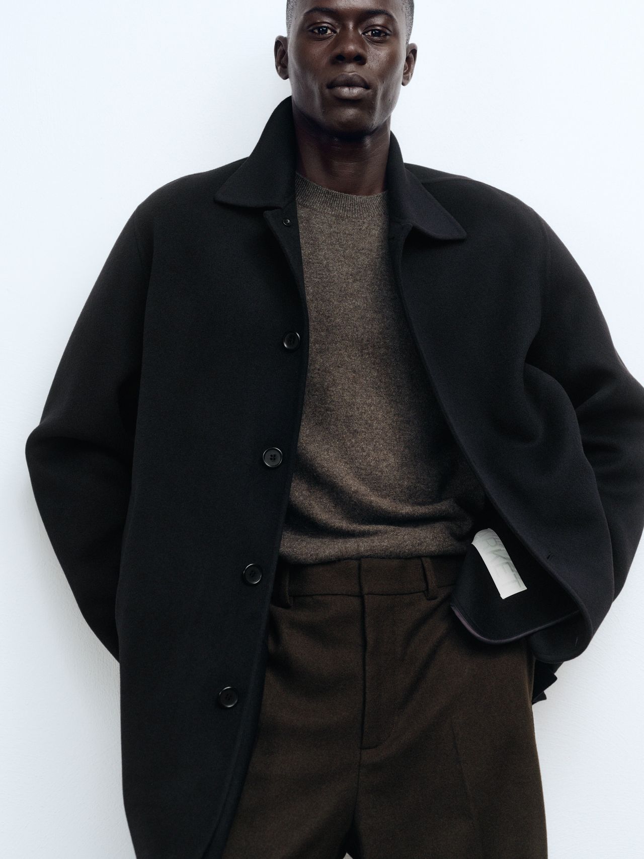 Person wearing a dark coat over a brown sweater and trousers, standing against a plain white background.