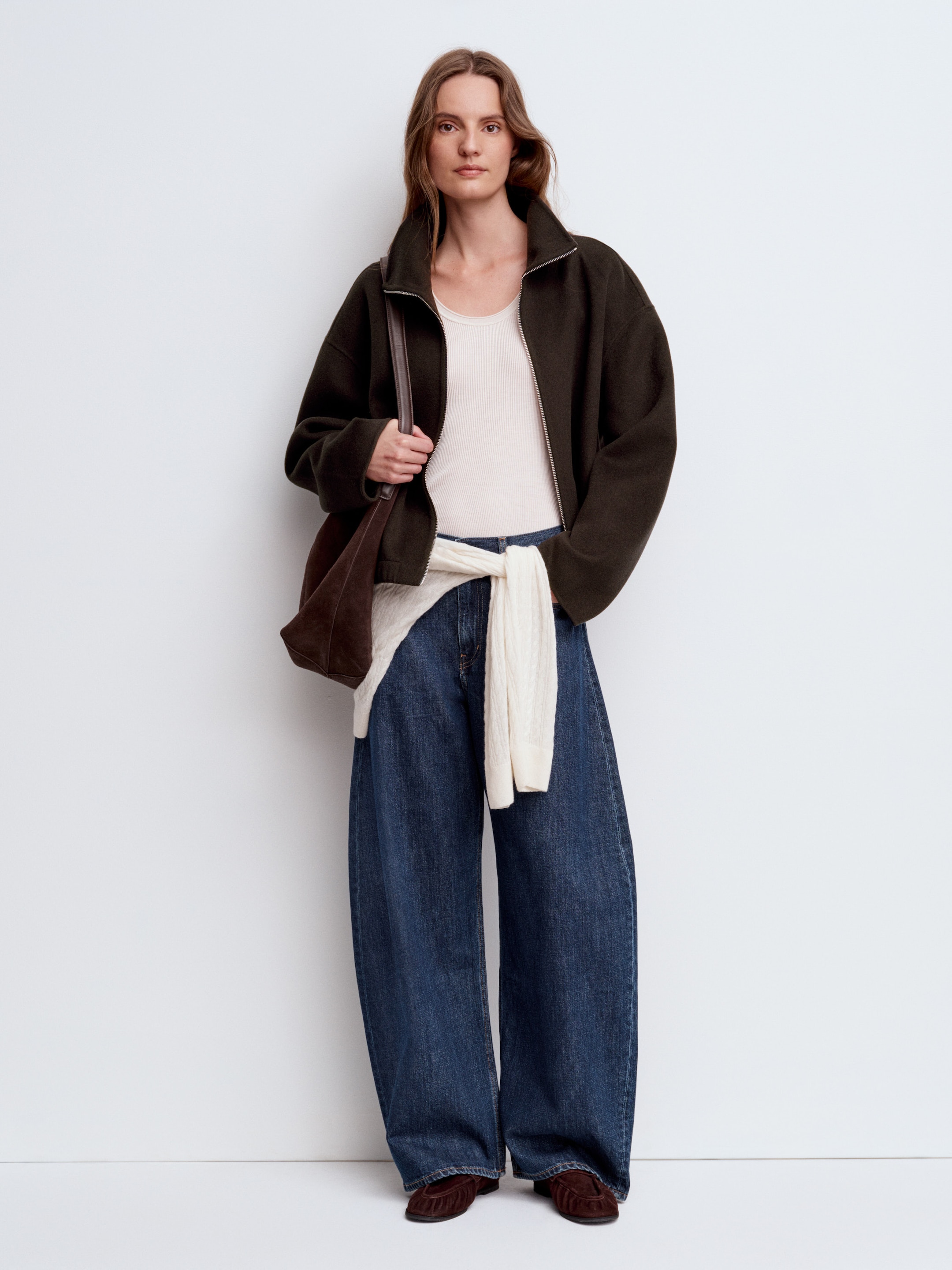 A person stands against a plain background, wearing a dark jacket, white top, wide-leg jeans, and holding a brown bag. A sweater is tied at the waist.